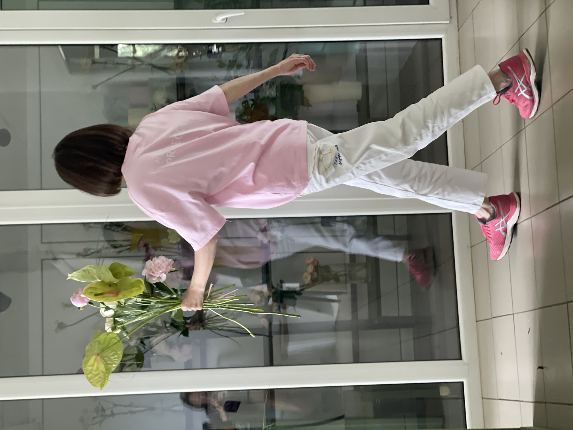 A woman in a pink t-shirt and white pants is holding a bouquet of flowers. She is standing in front of a glass door or window. The bouquet consists of pink peonies, green anthuriums, and some other foliage. The woman is wearing pink athletic shoes. The scene appears to be indoors, with tiled flooring visible. Reflections in the glass suggest an outdoor environment with greenery. The lighting is bright, indicating daytime. The overall mood is serene. There is text visible on the woman's pants, but it is too blurry to read.