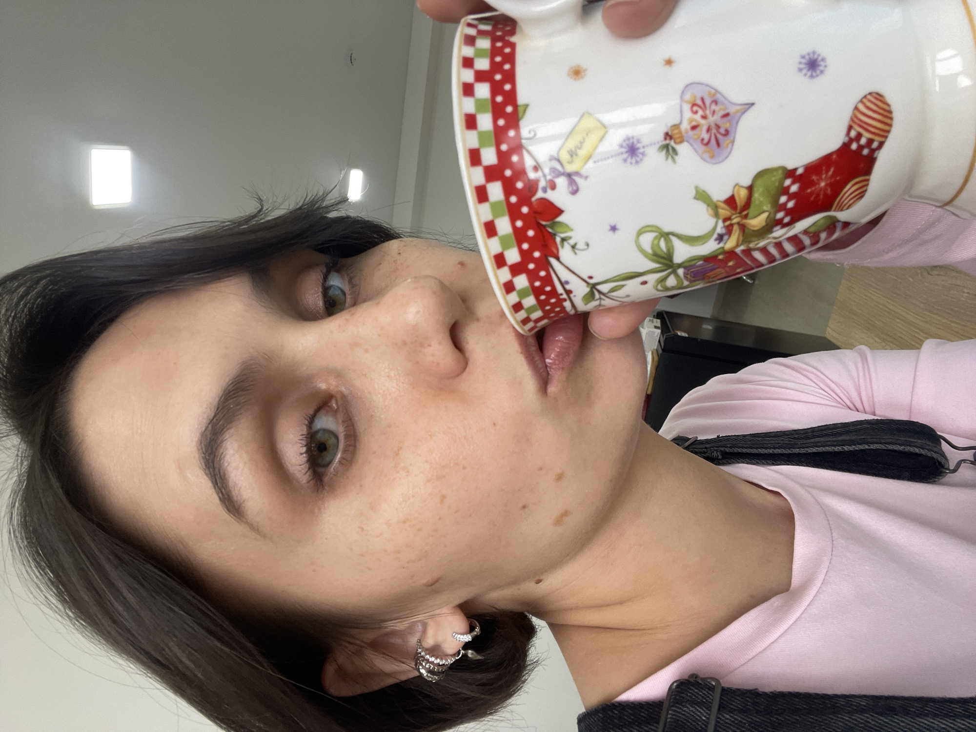 A young woman with short, dark hair and striking green eyes is captured in a close-up, rotated selfie, appearing to drink from a mug. Her face is speckled with freckles. She wears a light pink long-sleeved shirt, partially covered by dark overall straps, and prominent silver hoop earrings on her left ear. Her lips are touching the rim of a white ceramic mug, tilted upwards, revealing a glimpse of her tongue. Her left eye (viewer's right) is wide open and gazes towards the camera.
The mug is adorned with festive, Christmas-themed illustrations, including a red and white checkered pattern around the top rim, a red stocking with green ribbon and bow, decorative bells, stars, and snowflakes. A small yellow tag shape on the mug has faint, mostly illegible text, possibly reading "Nice" or "Noel."
The setting is indoors, characterized by bright, square ceiling lights visible in the upper background, suggesting a modern interior like an office or public building in Izhevsk, Russia. The background consists mainly of light-colored walls. The lighting is artificial and evenly distributed. The overall impression is a casual, candid moment.