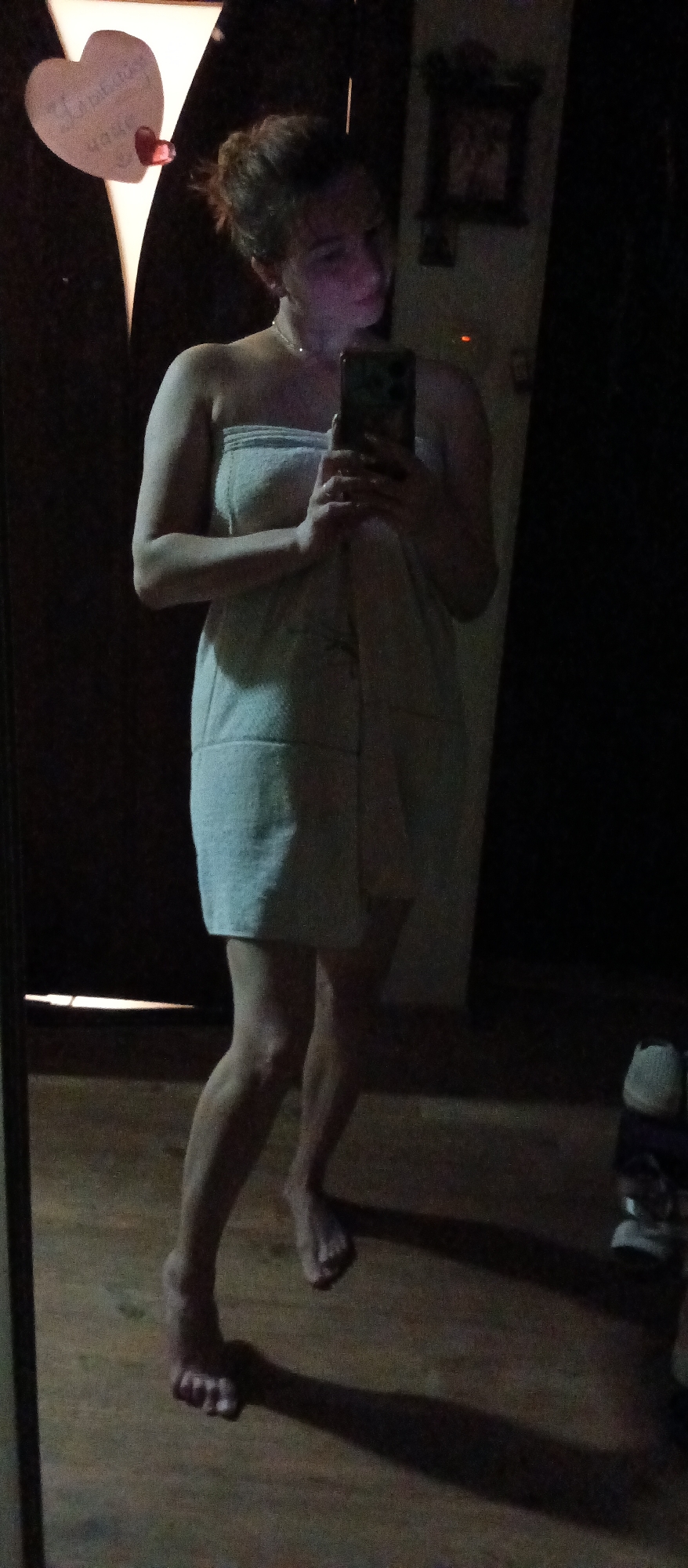A dimly lit, indoor scene shows a woman standing in front of a mirror, holding a mobile phone, presumably to take a selfie. She is wrapped in a white towel that covers her from chest to mid-thigh. Her hair is pulled back, and she appears to be barefoot. A necklace is visible around her neck.
Behind her is a dark wooden door adorned with a heart-shaped decoration featuring a handwritten message. A framed object is also visible on the wall in the background. The floor is made of wood. In the lower right corner, a pair of shoes is positioned on the floor. The setting appears to be a private residence, specifically a bathroom or bedroom. Location: Unknown, Unknown.