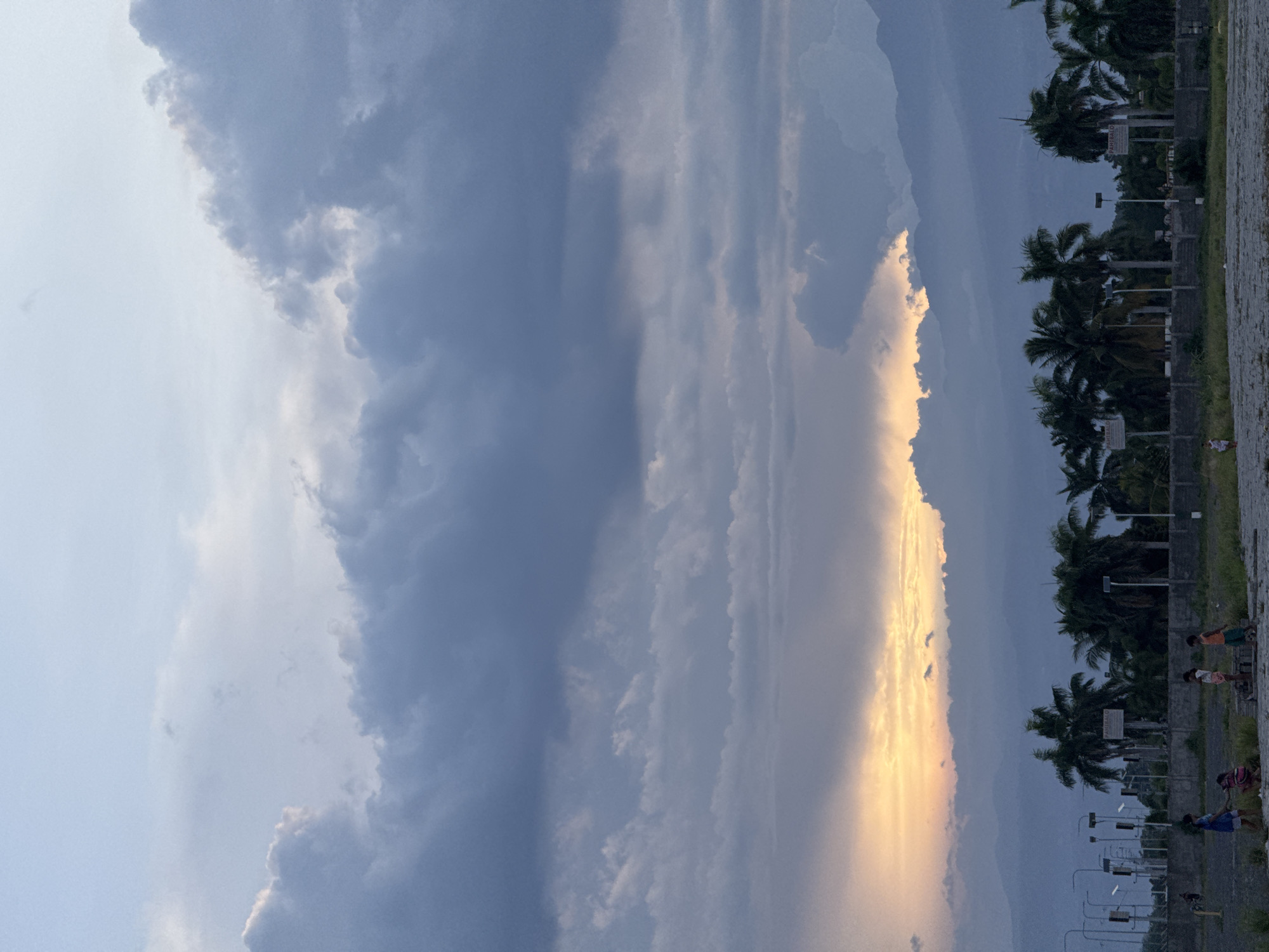 The image captures a scene in Davao City, Philippines, featuring a dramatic sky with dark, heavy clouds contrasting with a vertical column of bright, golden sunlight breaking through. In the lower portion of the image, along the right edge, there is a row of palm trees and what appears to be a paved or concrete walkway. Several people, including children, are present near the trees and walkway. There are also several light posts.