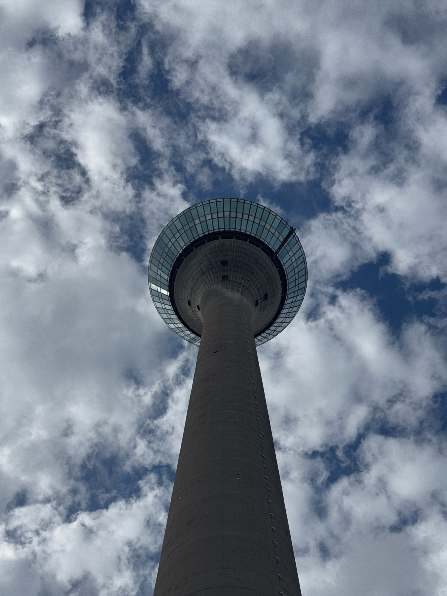 The image shows a low-angle view of the Rheinturm (Rhine Tower) in Dusseldorf, Germany. The tower, a concrete structure with a distinctive glass observation deck at the top, rises against a sky filled with scattered white clouds. The perspective emphasizes the height and imposing presence of the tower. The environment is urban, as indicated by the presence of this prominent landmark. There are no people visible in the image. The lighting suggests it is daytime, with diffused sunlight filtering through the clouds.