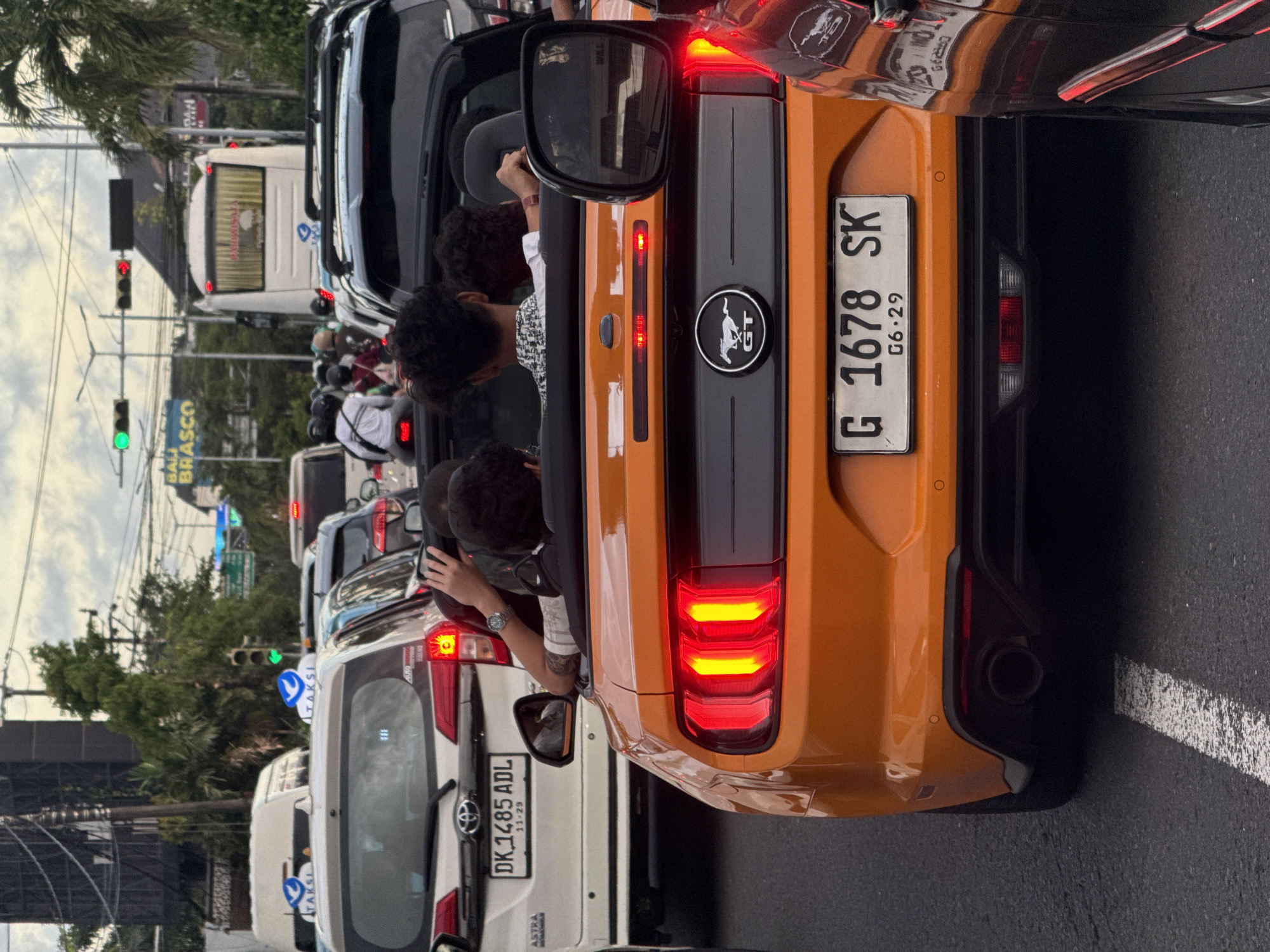 The image shows a traffic jam in Kuta, Indonesia. Several vehicles are stopped, including a white minivan taxi, and an orange Mustang convertible with several young male passengers. The Mustang's license plate reads "G 1678 SK" with a date code of "G6.29", and a 'GT' logo. The minivan taxi in front has a license plate of "DK 1485 ADL" with a date code of "11-29." A sign that says "BALI BRASCO" is visible in the background. The traffic light is green.