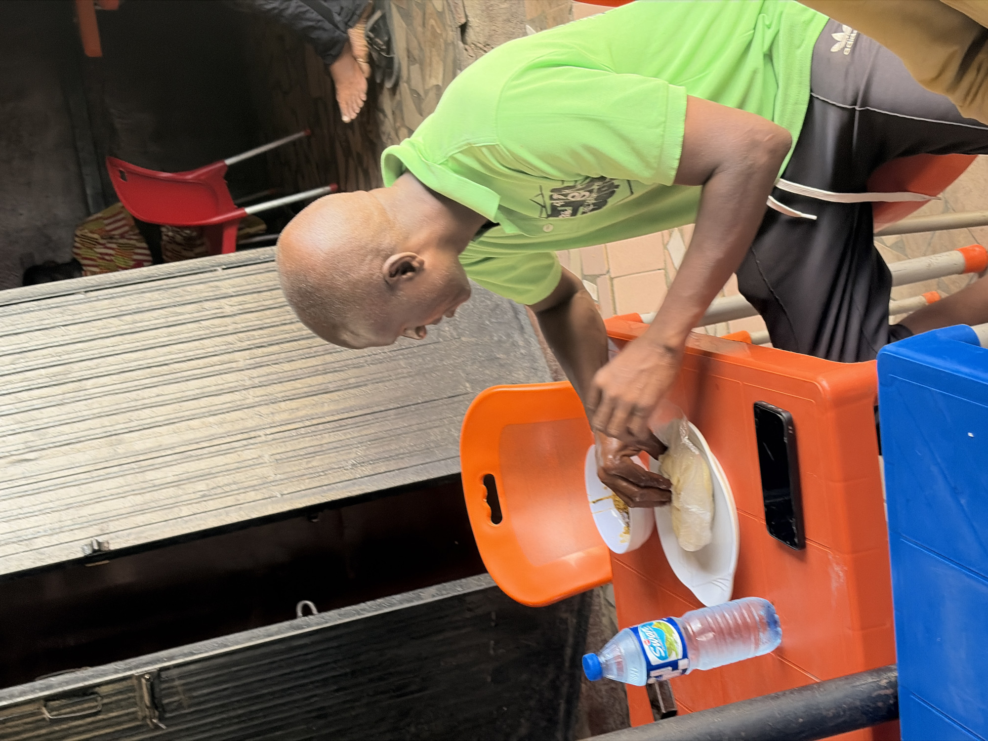 A person with dark skin and a bald head, wearing a bright lime green short-sleeved collared shirt and black athletic pants with an "ADIDAS" logo, is captured bent over, looking downwards. Their hands are positioned over a white plate holding a portion of food, which appears to be a light-colored, dough-like substance wrapped in plastic. A small white bowl with food remnants is also on the plate.
The person is leaning over an orange plastic surface, which seems to be a tray or part of a container. A black smartphone is propped vertically on this orange surface, and a plastic bottle of "SWAN Water" is lying horizontally nearby. To the right, part of a blue plastic container is visible.
In the left foreground, a prominent metallic or wooden slatted surface, possibly a counter or a top of a storage unit, spans horizontally. Below this, there's a dark, open space, suggesting a compartment or lower level. In the background, an overturned red plastic chair is visible on its side, next to a patterned fabric item. The wall behind has a textured, possibly tiled, appearance.
The scene appears to be an informal outdoor or semi-outdoor setting, possibly a market stall or a casual eating area. The lighting suggests it is daytime. The activity indicates the person is either preparing, unwrapping, or about to consume the food. The overall impression is a common daily life moment in Gura Top, Nigeria.