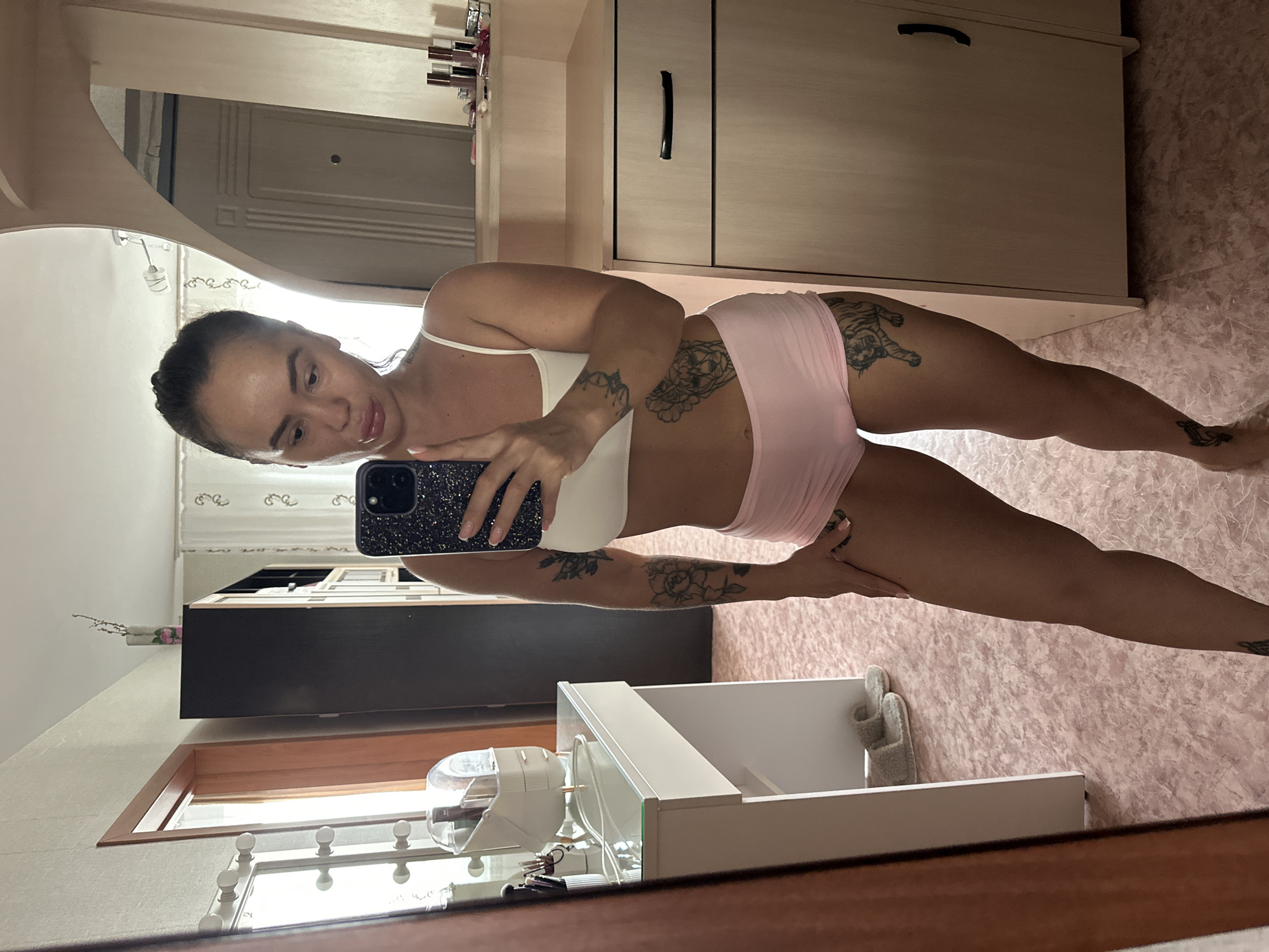 The image shows a woman taking a mirror selfie in a bedroom. She is wearing a white sports bra and pink shorts, and has several tattoos on her arms and legs. In the background, there is a vanity with makeup, a tall cabinet, and a closed door. The floor is covered in a pink patterned carpet, and a pair of slippers is visible near the vanity. The scene appears to be an indoor residential setting. The woman's phone is a dark color with a patterned case. There are decorative items on top of the cabinet, including what appears to be a vase with dried flowers and bottles of makeup or perfume. The lighting suggests it is daytime, with natural light coming from a window.