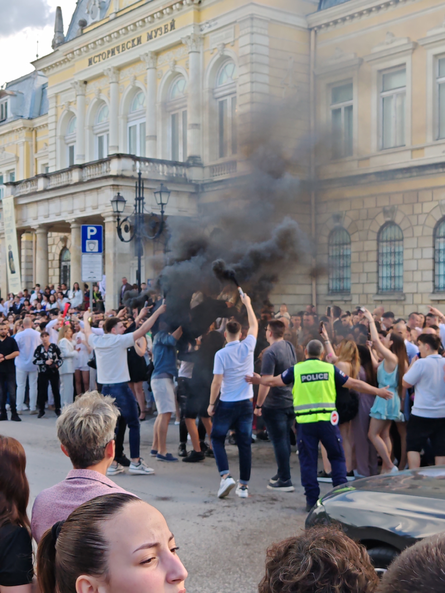 A medium eye-level shot shows a large crowd of people gathered in front of a building identified as the Historical Museum in Ruse, Bulgaria. In the foreground, a woman with her hair pulled back is visible from the chest up, looking to the left with a concerned expression. Behind her, a crowd of people, mostly young adults, are gathered. Several individuals are holding their arms up, and a plume of black smoke is rising from the center of the crowd. A police officer in a bright yellow vest with "POLICE" printed on the back is standing with his arms outstretched, seemingly attempting to control the crowd. In the background is the Historical Museum, a light-colored building with classical architectural features, including columns and arched windows. A "P" parking sign is visible in front of the building. The sky is overcast. A portion of a dark-colored vehicle is visible in the lower right corner.