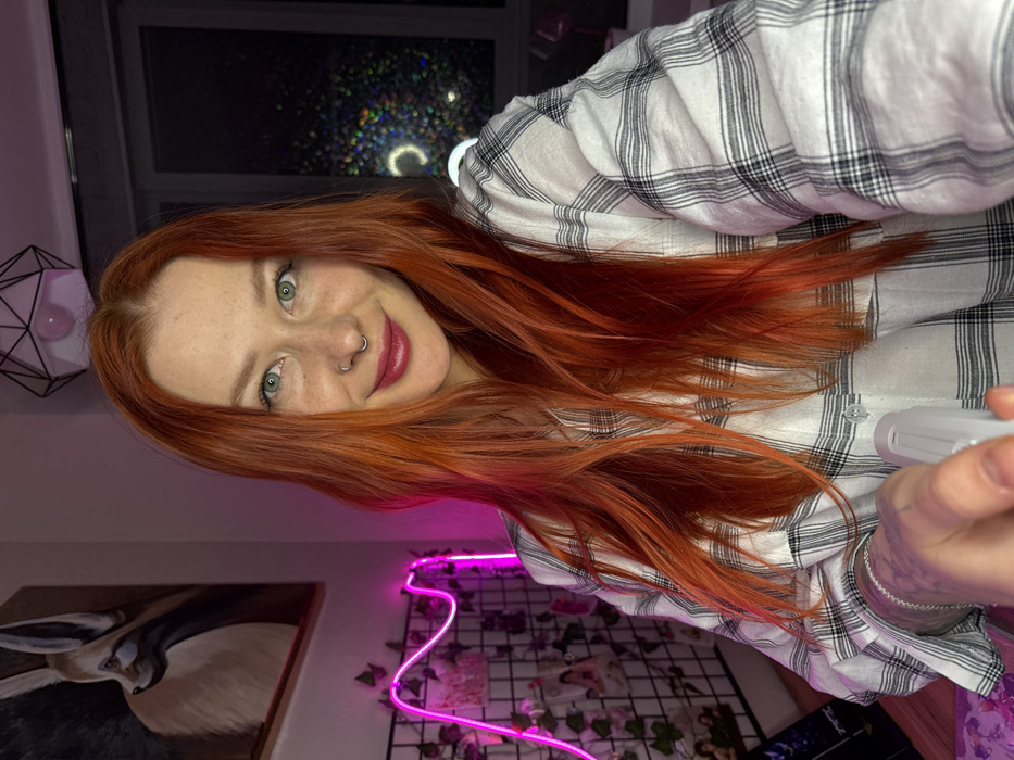 An indoor, close-up photograph depicts a female individual with long, wavy, auburn hair, freckles across her nose and cheeks, and green eyes. She wears dark red lipstick, a silver septum piercing, and a silver stud in her left nostril. Her attire consists of a white and black plaid button-up shirt, with the left sleeve revealing a tattooed forearm. She is positioned centrally, looking directly at the viewer with a slight smile. Her left hand extends forward, holding a translucent white object, possibly a spray bottle or similar device, pointed towards the camera's perspective.
The background to her left features a framed artwork depicting a Fennec fox in profile against a dark background, positioned above a black grid wall panel. A pink undulating neon light is mounted on or near this grid, intertwined with artificial green foliage. Multiple small photos or notes are affixed to the grid. To her right, a window shows a dark exterior, with prominent circular and speckled light reflections visible on the glass, including what appears to be a ring light reflection. A white geometric pendant light fixture with an exposed bulb is suspended from the ceiling above her head. The scene is set in an interior space within the city of Rostov-on-Don, Russia.