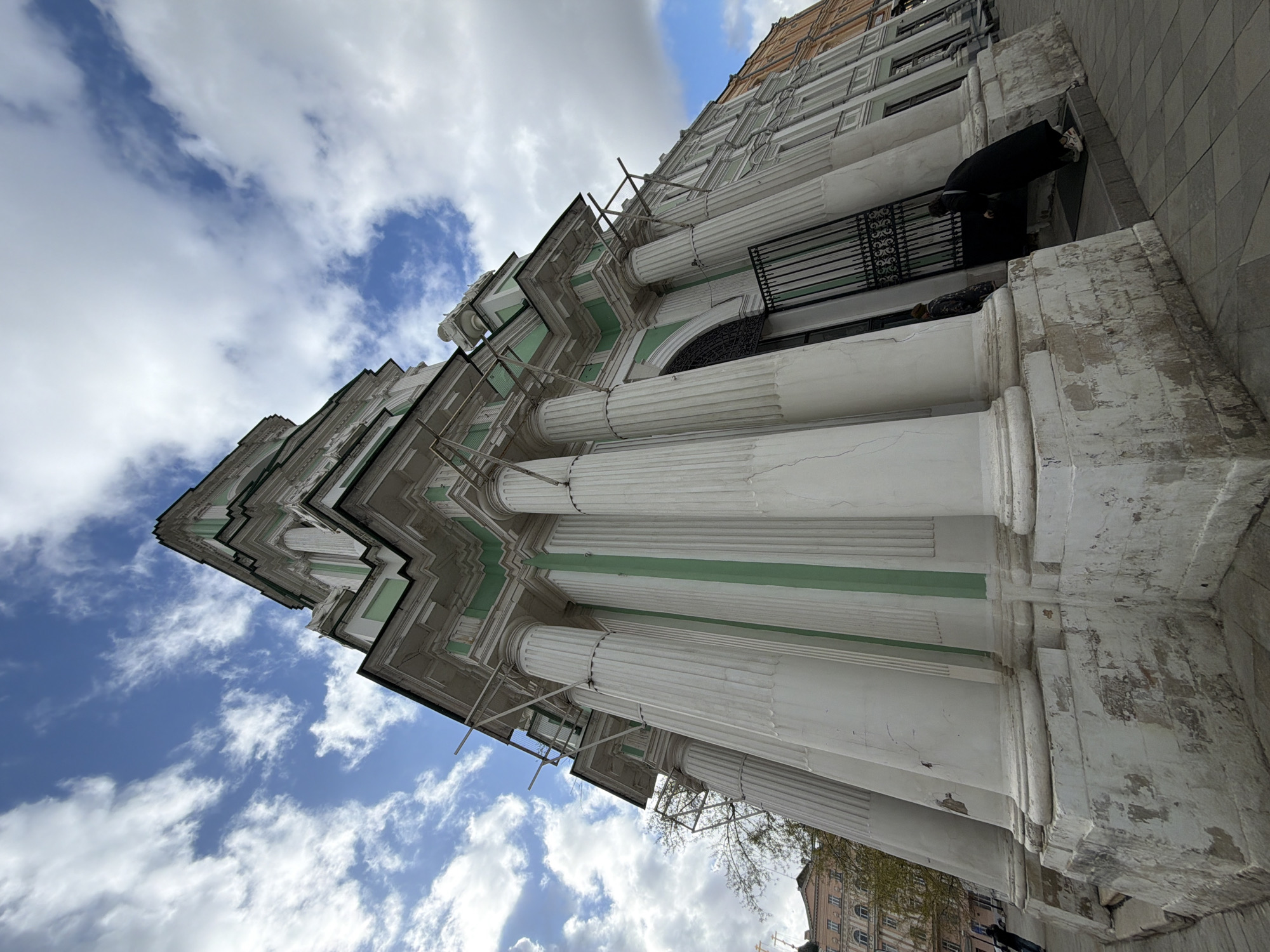The image depicts the facade of a classical building with white columns and green accents against a backdrop of a partly cloudy sky in Moscow, Russia. Scaffolding is present on the building, indicating ongoing maintenance or restoration work. A person in dark clothing is standing on a balcony or ledge of the building. The perspective is from a low angle, emphasizing the height and grandeur of the architecture.