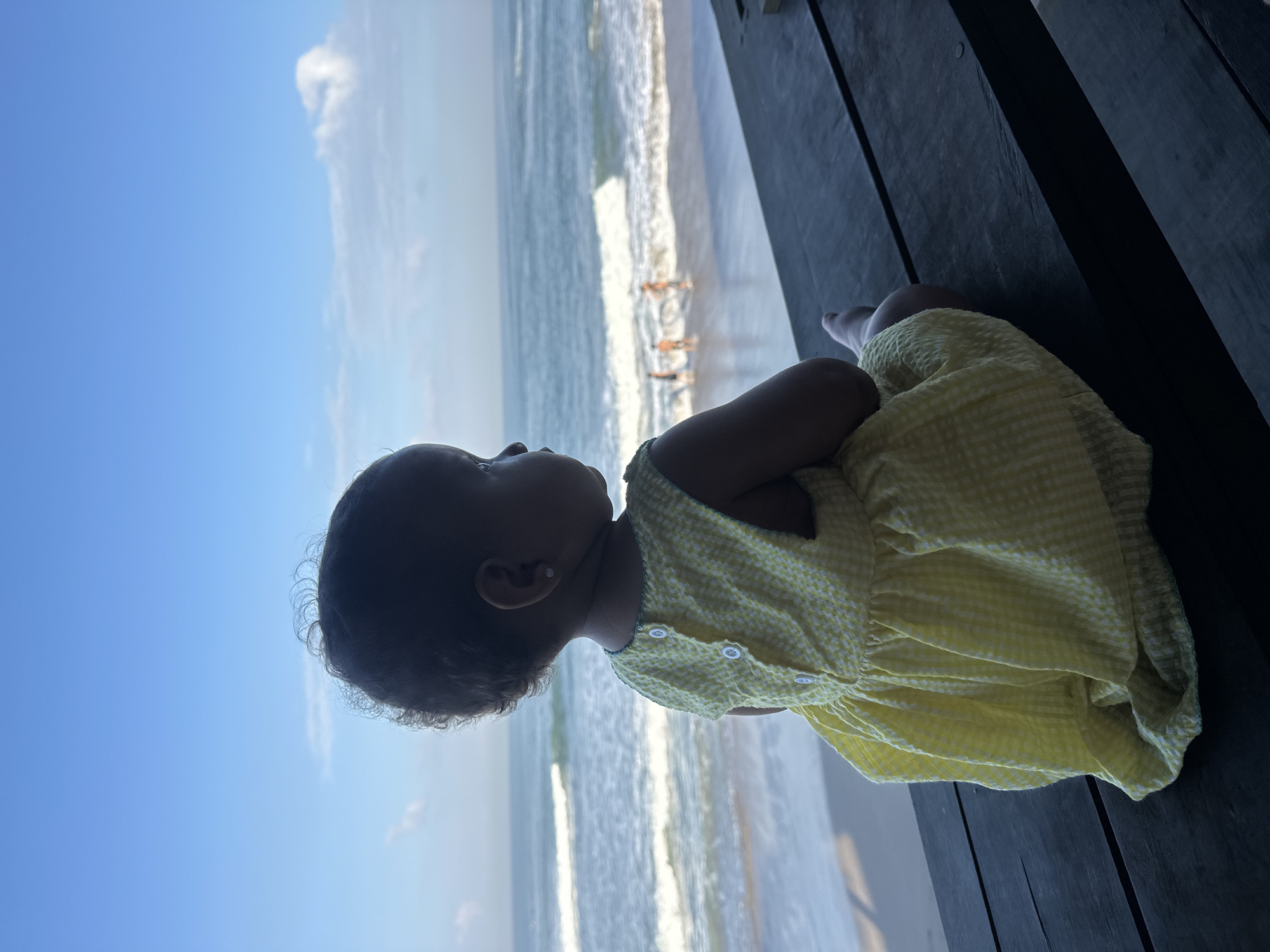 An infant, facing away from the camera, sits on a dark wooden surface, likely a table or bench, and gazes out at the ocean. The infant is wearing a yellow gingham dress. In the distance, on the beach and in the water, several people can be seen, appearing small due to the distance. The scene suggests a coastal location, possibly in North Kuta, Indonesia, characterized by a clear blue sky with a few scattered clouds, and gentle waves rolling onto the shore.