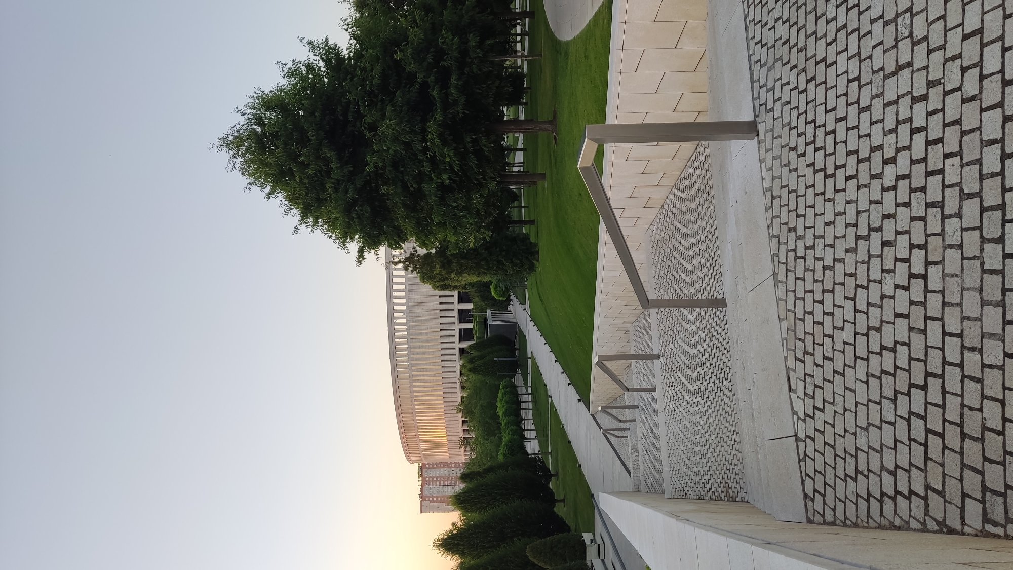 The image displays a modern architectural complex situated in an outdoor park-like setting. The dominant structure is a large, curved building with a white, ribbed facade, suggesting it might be a stadium or a convention center. In the foreground, a paved walkway made of small, light-colored cobblestones runs parallel to a manicured lawn. A metal railing runs along the edge of the walkway, guiding visitors.
Scattered throughout the green expanse are various trees, some tall and mature with dense foliage, others smaller and more stylized, forming a decorative border. In the distance, a few residential buildings can be seen, indicating the complex is likely within or near a city.
The sky is a soft, pale blue with a hint of orange at the horizon, suggesting it is either early morning or late evening, during the golden hour. The lighting is gentle, casting soft shadows and highlighting the clean lines of the architecture and the vibrant green of the grass. The overall impression is one of serenity and modern design seamlessly integrated with nature. There is no visible text in the image. The scene appears to be located in Krasnodar, Russia, given the provided context.