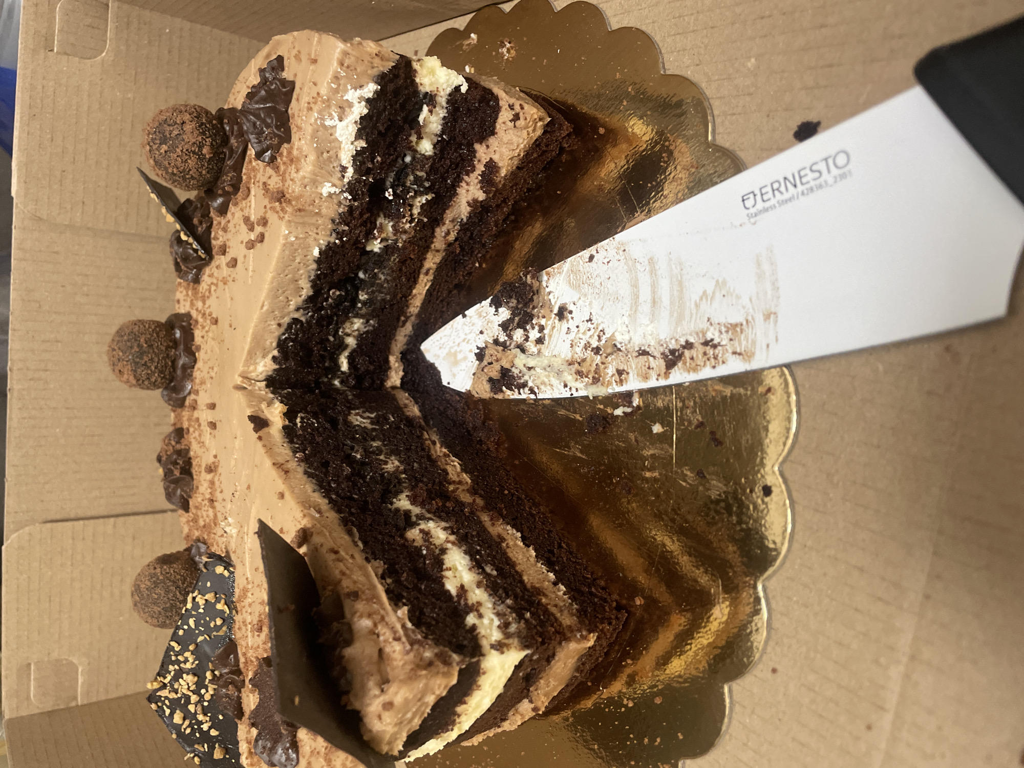 A partly consumed chocolate cake with multiple layers of chocolate cake and light-colored cream is inside a cardboard box. A knife with the brand "ERNESTO" printed on its blade is cutting into the cake. The cake is decorated with chocolate truffles and dark chocolate shavings. The cake is sitting on a gold-colored scalloped tray. The scene is likely indoors. The location in Sofia, Bulgaria cannot be determined.