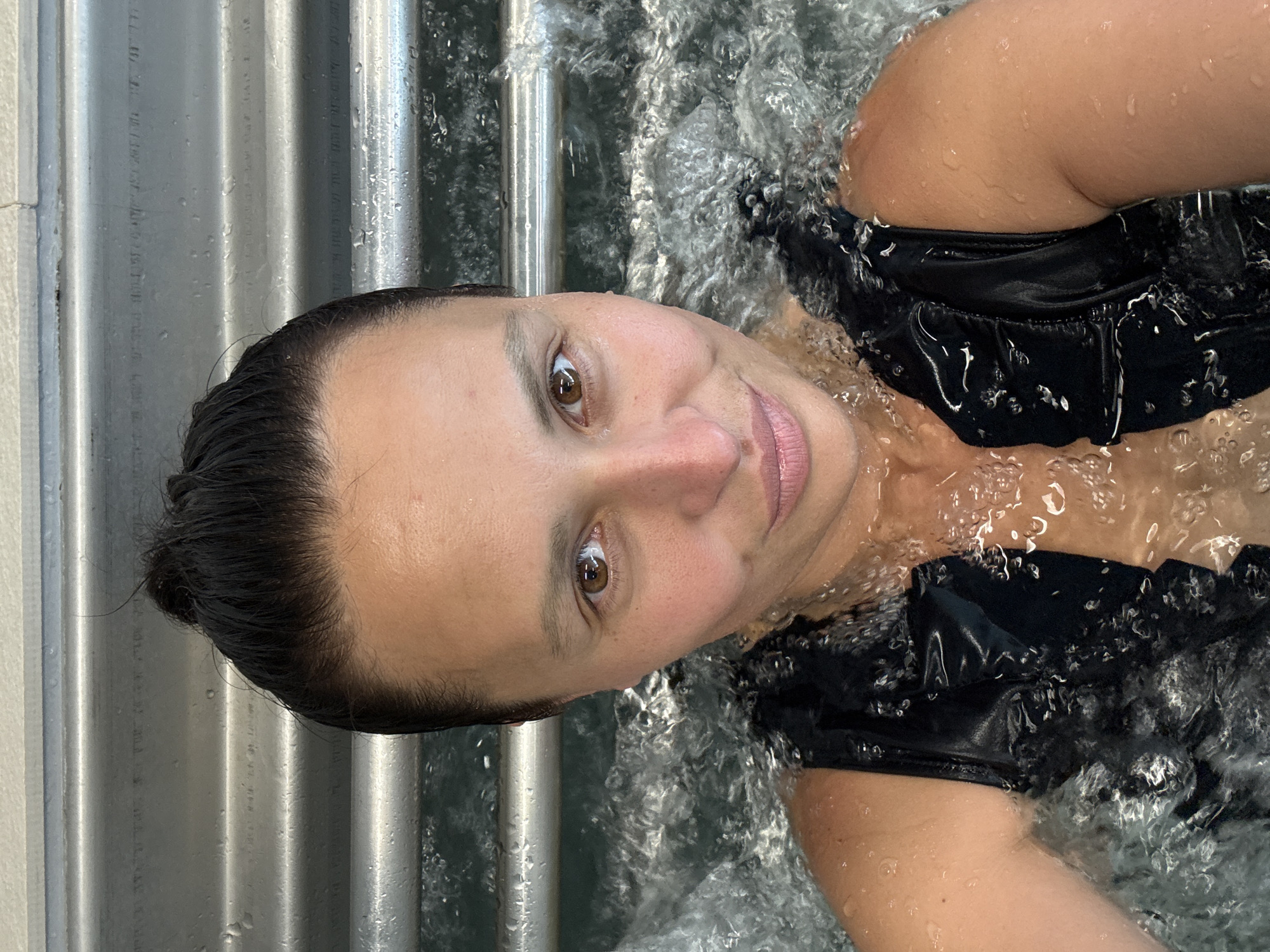 The image is a close-up, high-angle selfie of an adult female partially submerged in bubbling water, consistent with a hot tub or jacuzzi. She has dark hair pulled back, and her brown eyes are open, looking directly at the camera with a subtle smile. Water droplets are visible on her face, neck, and shoulders, and she wears a dark, possibly black, swimsuit or top. The water around her torso is vigorously agitated with numerous white bubbles and splashes. The background behind her head features a metallic, horizontally ribbed wall, likely the interior rim of the tub, with a lighter-colored, possibly tiled, surface visible above it at the top of the frame.