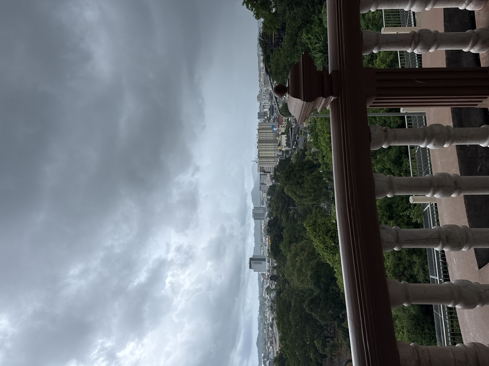 The image depicts a cityscape under a cloudy, overcast sky, likely taken from a balcony or elevated vantage point in Phuket City Municipality, Thailand. In the foreground, a decorative balustrade made of white balusters and a dark brown railing frames the view. Beyond the railing, a dense expanse of lush green trees dominates the mid-ground, transitioning into a city skyline with various buildings, including taller apartment complexes and office buildings. Mountains are visible in the distance, softened by the hazy atmosphere. The sky is filled with heavy, grey clouds, suggesting impending or ongoing rain. There are no people or visible actions in the scene. The overall impression is one of a tranquil, slightly moody urban environment surrounded by nature.