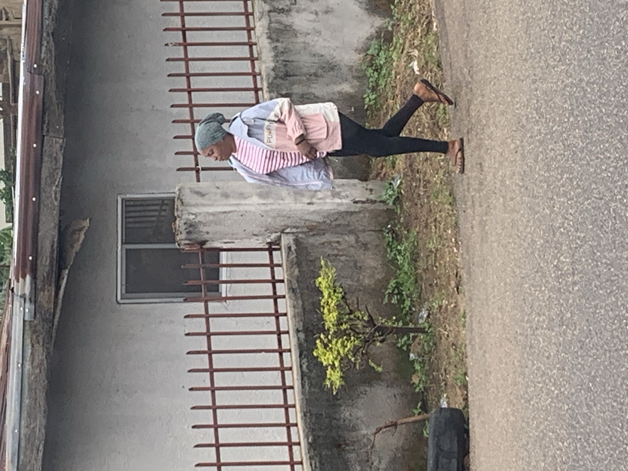 The image depicts a daytime scene in what appears to be a semi-urban or residential area, likely in Jos, Nigeria, given the provided context.
The primary subject is a person, seemingly a woman, standing on the unpaved shoulder beside a grey asphalt road. She is dressed in a light grey head covering, a light-colored long-sleeved top (possibly blue and white) worn over a pink and white striped shirt, black leggings, and brown flip-flops. Her head is tilted slightly downwards, and she is looking away from the camera.
Behind the person, to the left, is a building with a textured grey concrete wall. A small window with security bars is visible on this wall. A brown metal fence runs across the mid-ground, separating the building from the foreground. On the far left, a section of a roof with rusty corrugated metal sheeting can be seen.
Between the fence and the road, there is a strip of ground with sparse green vegetation, including a small bush or tree with bright green leaves. A dark, possibly discarded, tire is also visible near the edge of the road. The road itself is paved with a granular grey material.
The lighting suggests it is daytime, with no strong shadows indicating early morning or late afternoon. The scene captures an ordinary moment, with no specific activity beyond standing or pausing. On the striped shirt worn by the person, partial text "PLUM..." is discernible.