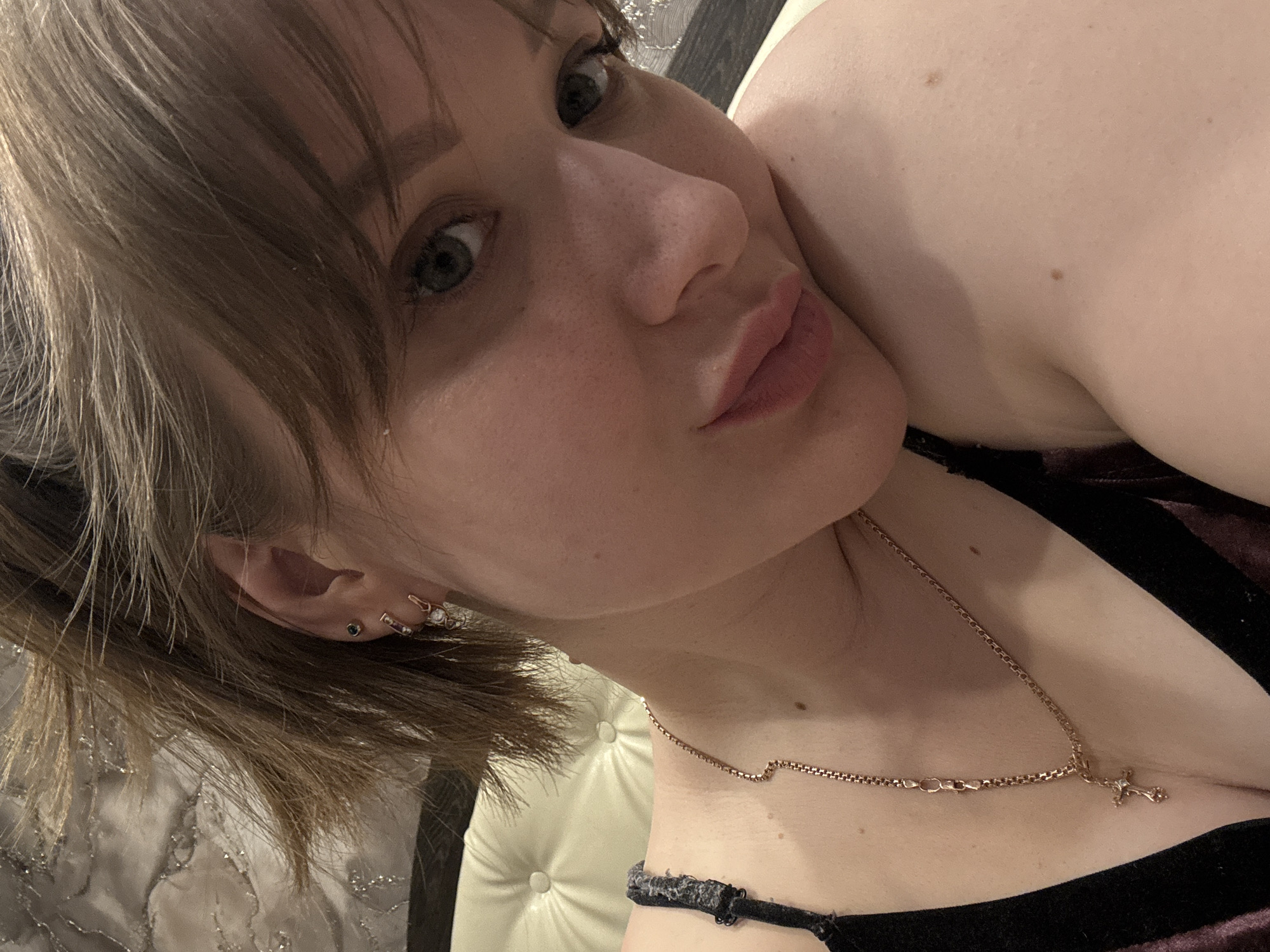A close-up, slightly angled selfie of a fair-skinned woman with short, light brown hair. She has light-colored eyes and is wearing earrings and a delicate gold necklace with a cross pendant. She is wearing a dark-colored top with a visible strap. The background is blurred and includes a tufted light-colored headboard. The location cannot be determined as Chaikovsky, Russia based solely on the information in the image.
