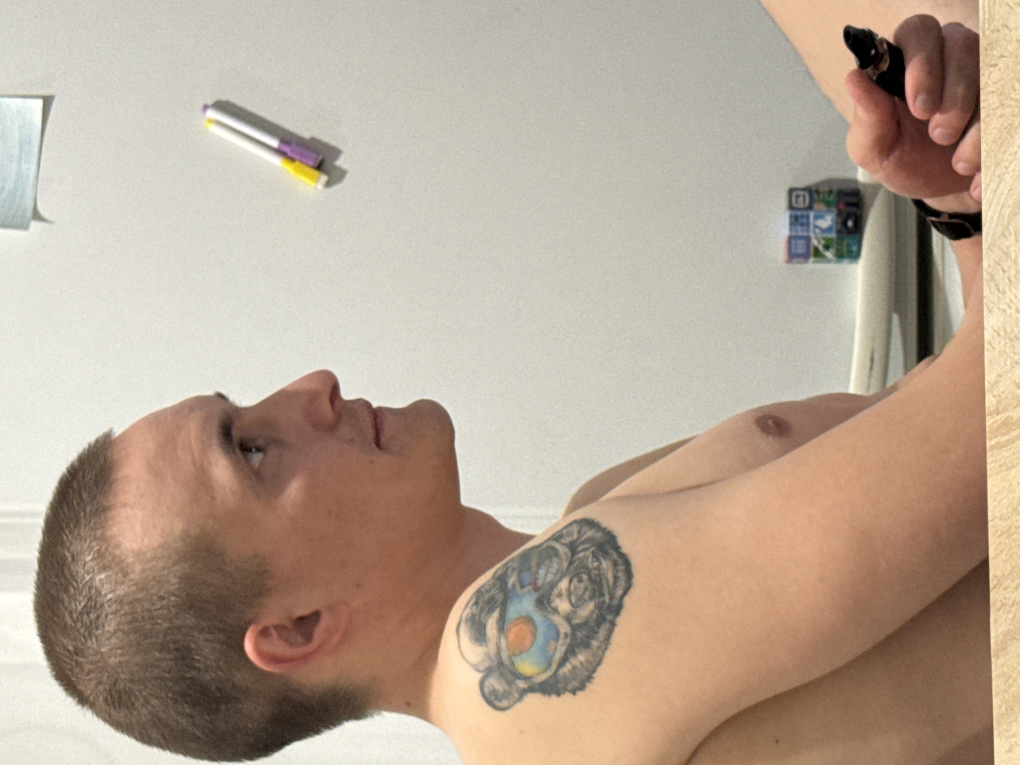 A shirtless man with a short haircut and a tattoo on his left shoulder is looking up and to the left. In his right hand, which is above his head, he holds a black object. On the wall behind him, there is a sticky note, two markers, and a small, square object with multiple images. There is no indication in the photo that this image was taken in Chaikovsky, Russia.