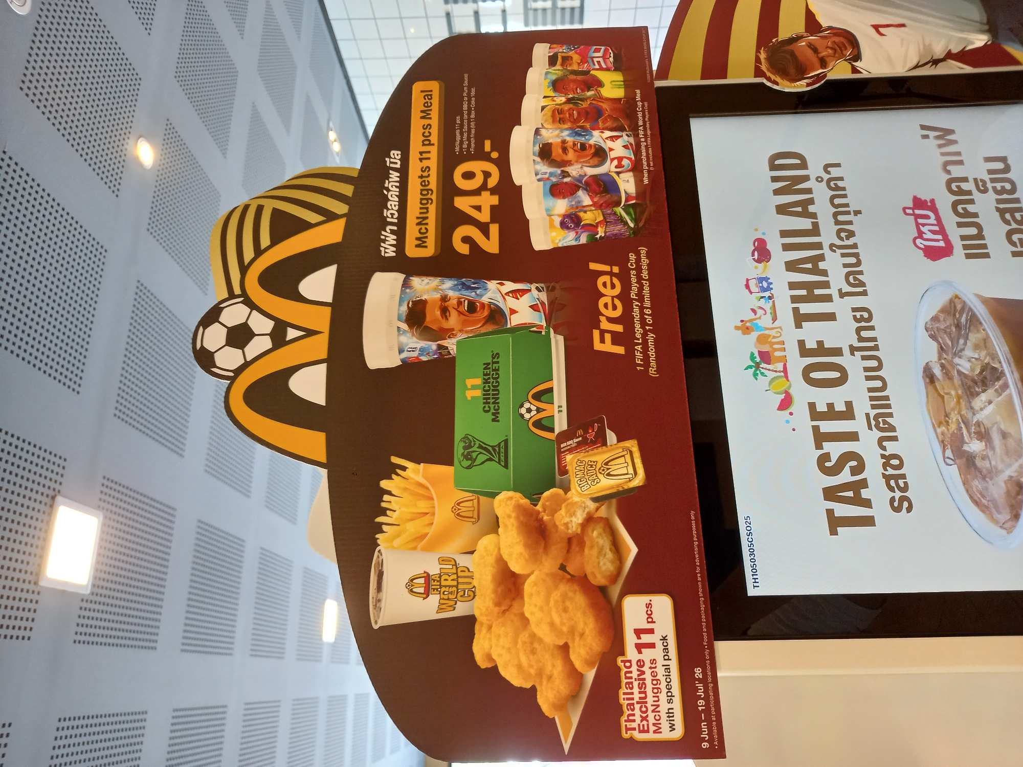 This is a medium shot, eye-level photograph of a promotional display for McDonald's in Baan Bo Phut, Thailand. The display features a large, curved banner with a dark red background and a wavy yellow and black pattern at the top, accented by a soccer ball. In the foreground, a pile of 11 McNuggets is arranged on a yellow paper liner. To the left, a text box reads "Thailand Exclusive 11 pcs. McNuggets with special pack."
Behind the McNuggets, several McDonald's branded items are visible. A white cup with "FIFA WORLD CUP" printed on it and a cup of french fries are prominently displayed. A green box with the FIFA World Cup logo and "11 CHICKEN MCNUGGETS" is also present. Small packets of sauce are next to the McNuggets, one labeled "BIG MAC SAUCE".
To the right of the McNuggets, a white cup with a soccer player's portrait is displayed, and below it, a row of six smaller cups, each featuring different soccer player portraits, are shown. Text on this side of the display indicates "McNuggets 11 pcs Meal" and lists the meal's contents and price: "249.-". A section below that reads "Free! 1 FIFA Legendary Players Cup (Randomly 1 of 6 limited designs)".
At the bottom of the frame, a digital screen displays "TASTE OF THAILAND" in large English letters, followed by Thai script. A partial image of an iced beverage is visible on the screen. The lighting appears to be from overhead fluorescent lights, common in indoor retail environments. The overall impression is a vibrant and enticing advertisement for a limited-time McDonald's promotion.