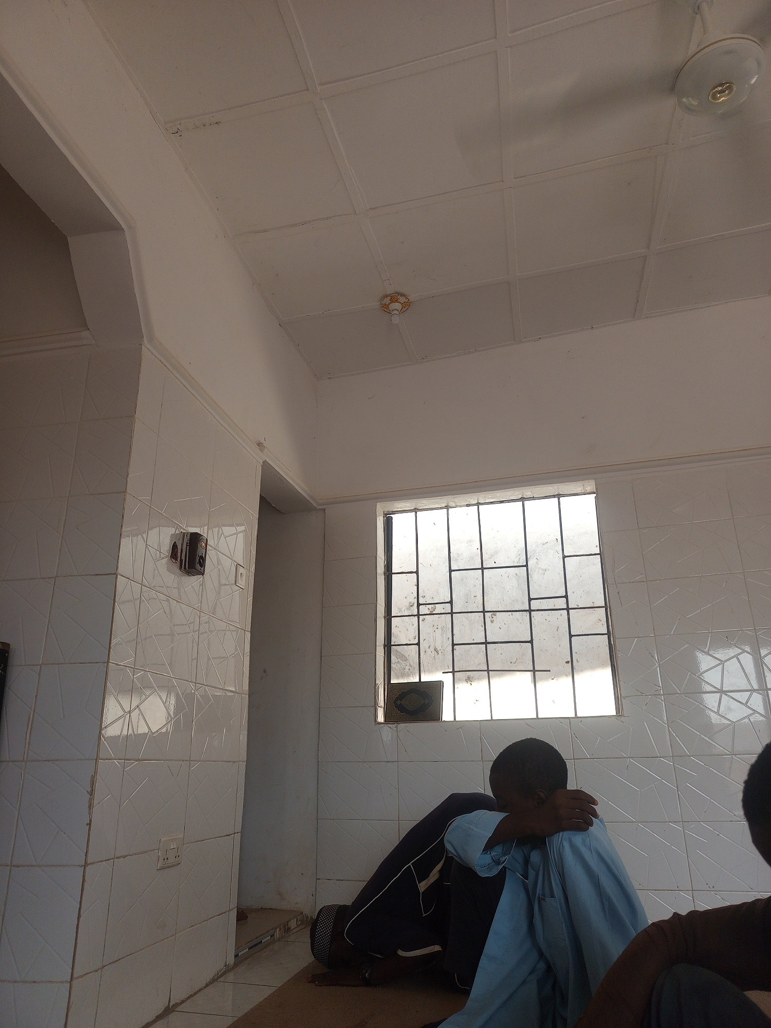 The image displays an interior room in Anguwar Jaba, Nigeria, with white tiled walls featuring a subtle geometric relief pattern and a white panelled ceiling. Two ceiling-mounted light fixtures are present: a central white ceiling fan with an integrated light, and a smaller, ornate gold and white lamp near the upper right corner.
A rectangular window, secured by a dark metal grid, is visible on the back wall, admitting significant natural light through its dirty panes. A small, dark, possibly decorative or religious item rests beneath the window. Electrical components are installed on the tiled wall to the left: a dark, rectangular control unit with a circular dial, and a white duplex electrical outlet below it. A smaller white switch or outlet is positioned to the right of the control unit. A decorative white molding runs horizontally along the top edge of the tiled walls.
The floor is covered by a light brown mat or rug. In the mid-ground, a young male individual with dark skin and short hair is seated on the mat, facing towards the left. He wears a light blue long-sleeved garment over dark trousers, and his right arm is bent, with his hand near his head, obscuring his face in a posture of rest or contemplation. Dark footwear, possibly sandals, is visible near his feet. Partially visible in the immediate foreground, on the right, is the upper back of another individual wearing a brown top and dark trousers. A doorway or archway to the left of the window reveals a darker area beyond.