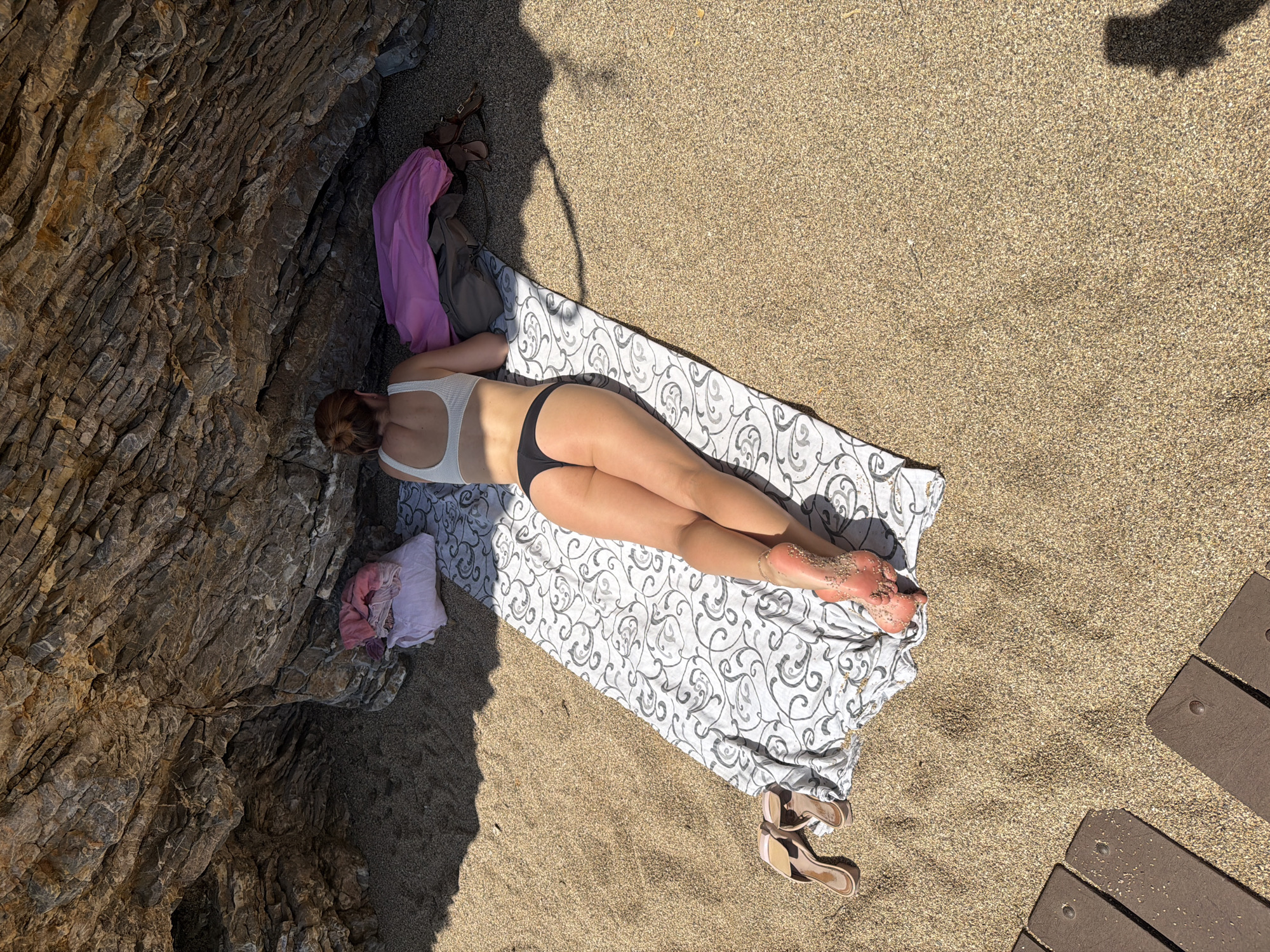 The image captures a woman sunbathing on a beach in Budva, Montenegro. She is lying face down on a patterned towel, wearing a white top and black bikini bottoms. Her feet are covered in sand. To her left, a large rock formation provides shade, and items like clothes and a bag are placed near it. A pair of sandals are beside the towel. The rest of the scene consists of sandy beach and a section of wooden walkway.