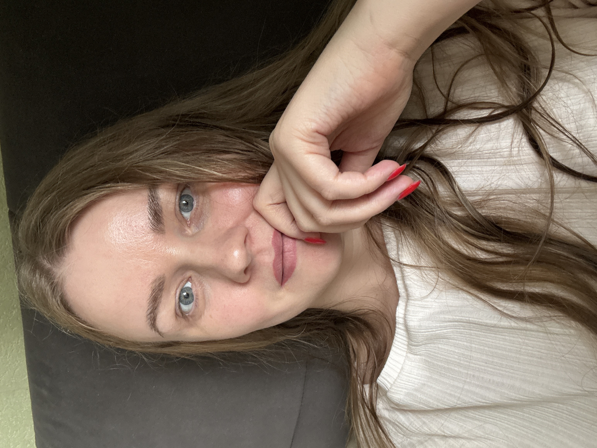 A young woman lies supine on what appears to be a dark gray or black couch. She has light skin, long brown hair, and blue eyes. Her left hand rests on her face, with bright red nail polish on her fingernails. She is wearing a white top. The background is out of focus. There is no definitive information to confirm the location is Chaikovsky, Russia.