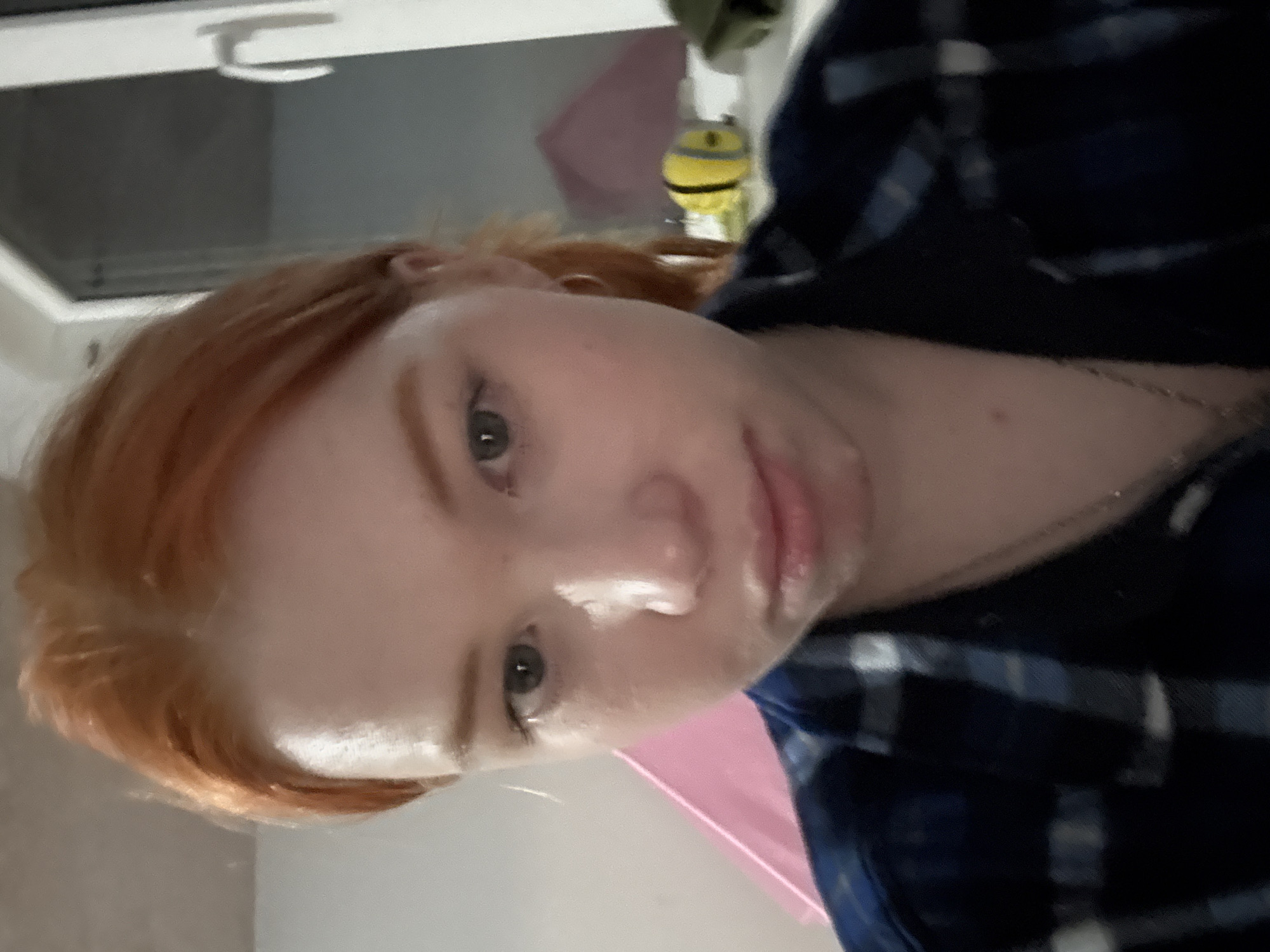 The image features a young individual with reddish-blonde hair and light green or hazel eyes, captured in a close-up selfie that is rotated 90 degrees clockwise. The subject is looking directly into the camera with a neutral expression, their lips appearing slightly glossy. They are wearing a dark blue shirt with a light-colored plaid pattern and a thin, silver-toned chain around their neck.
The setting appears to be indoors. In the background, to the subject's left, a white-framed window or skylight with a dark pane is visible. To the right, behind the subject's shoulder, there's a small, yellow object resembling a Minion character, and a soft pink object, possibly a piece of furniture or decor. The lighting creates highlights on the subject's forehead and lips. No specific activity beyond posing for a photo is discernible. There is no visible text in the image.