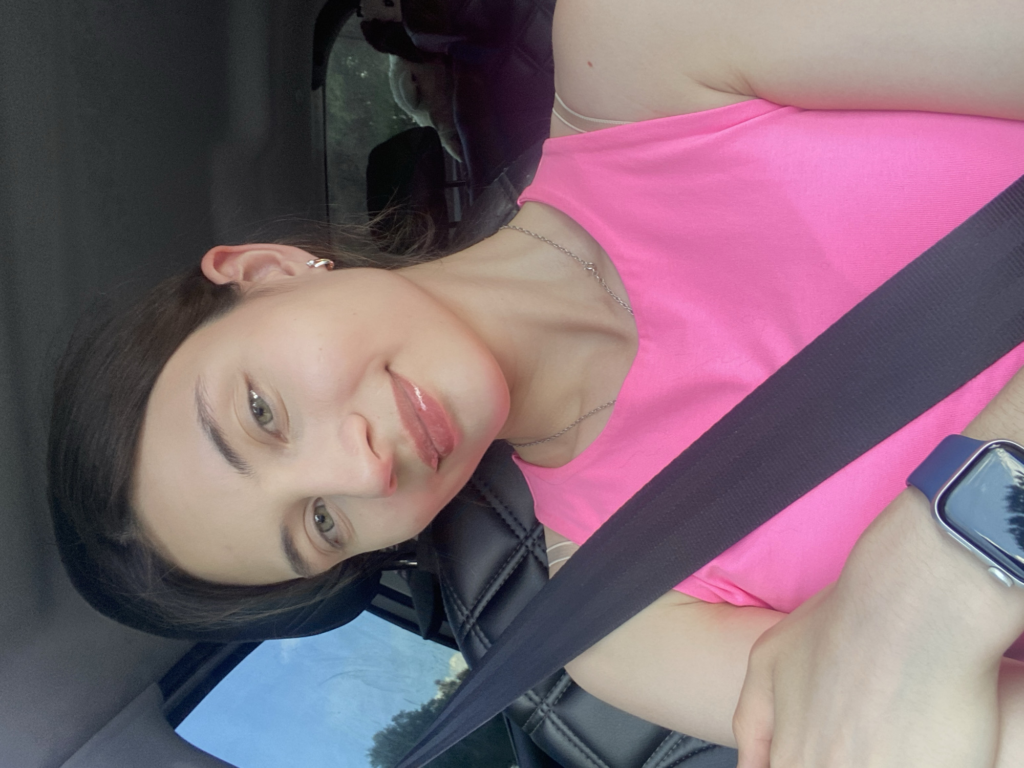 A young woman with light brown hair and green eyes is shown in a selfie taken from inside a car. She is wearing a bright pink tank top, a silver necklace, and hoop earrings. A black seatbelt crosses her chest. On her left wrist, she wears a blue smartwatch. The background shows the interior of the car, including a black quilted seat and a window with a view of trees and the sky. In the backseat, a fluffy white dog is visible. The image was taken in the city of Kamkoye, Russia.