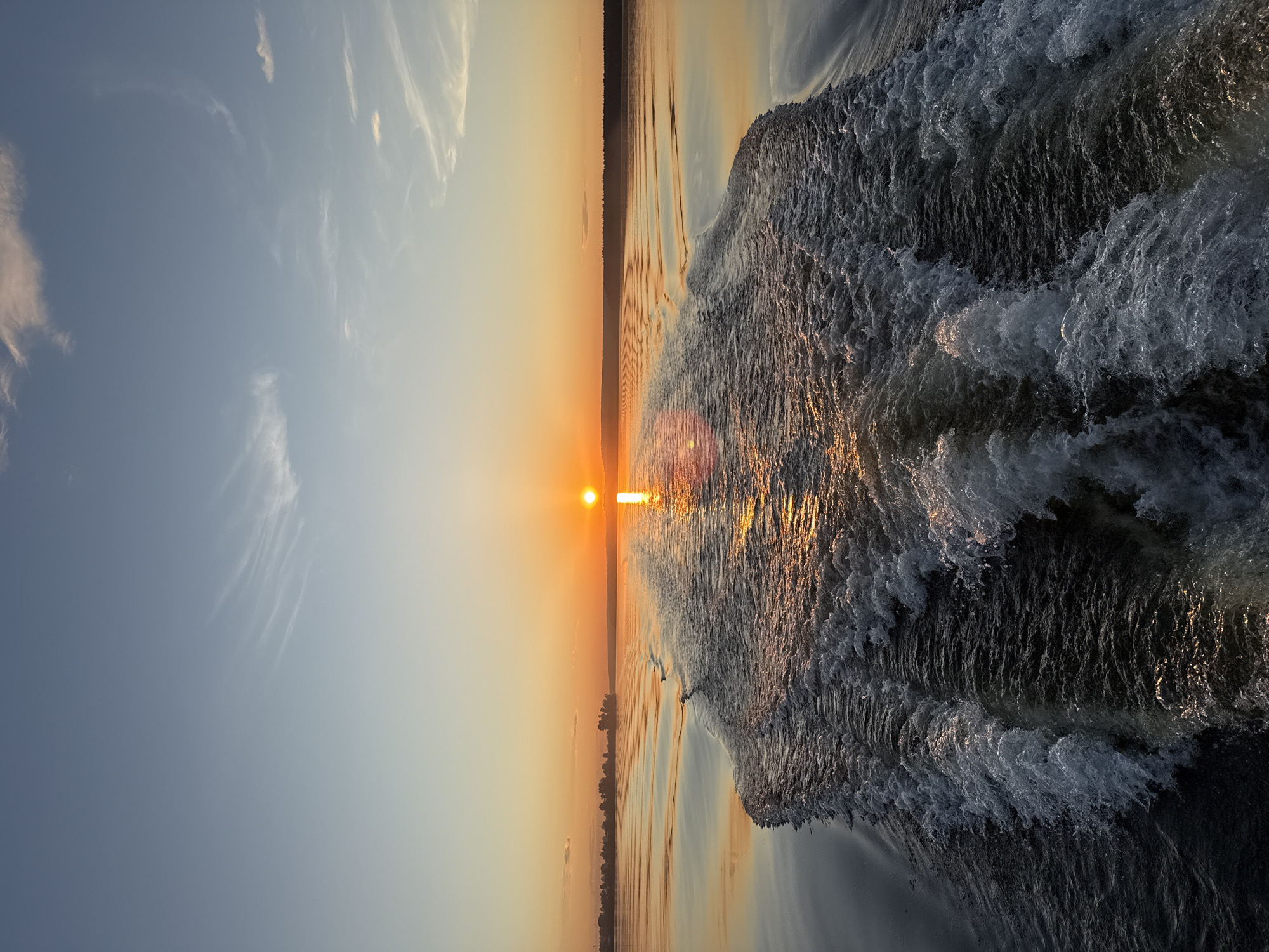 This image captures a wide-angle view of a sunset or sunrise over a large body of water, likely the Votkinsk Reservoir in Votkinsk, Russia. The perspective is from the stern of a moving vessel, evidenced by the turbulent wake extending from the foreground to the horizon.
The sky in the upper portion is a clear, pale blue with faint, wispy cirrus clouds scattered across it. This blue transitions downwards into a warm, luminous orange-yellow at the horizon. The sun, a bright, intense orange-yellow disc, is centered on the horizon, creating a prominent glare and an elongated reflection path across the water directly in front of it.
The water features a wide, frothy wake composed of churned white water, indicating a boat's recent passage. This wake forms a central channel leading towards the sun's reflection. On either side of the wake, the water is calmer, reflecting the golden and orange hues of the sky and the sun. Distant landforms, comprising dark silhouettes of trees and possibly low-lying structures, line the horizon to the left and right, framing the central water expanse. No individuals or specific man-made objects other than the implied vessel creating the wake are discernible.