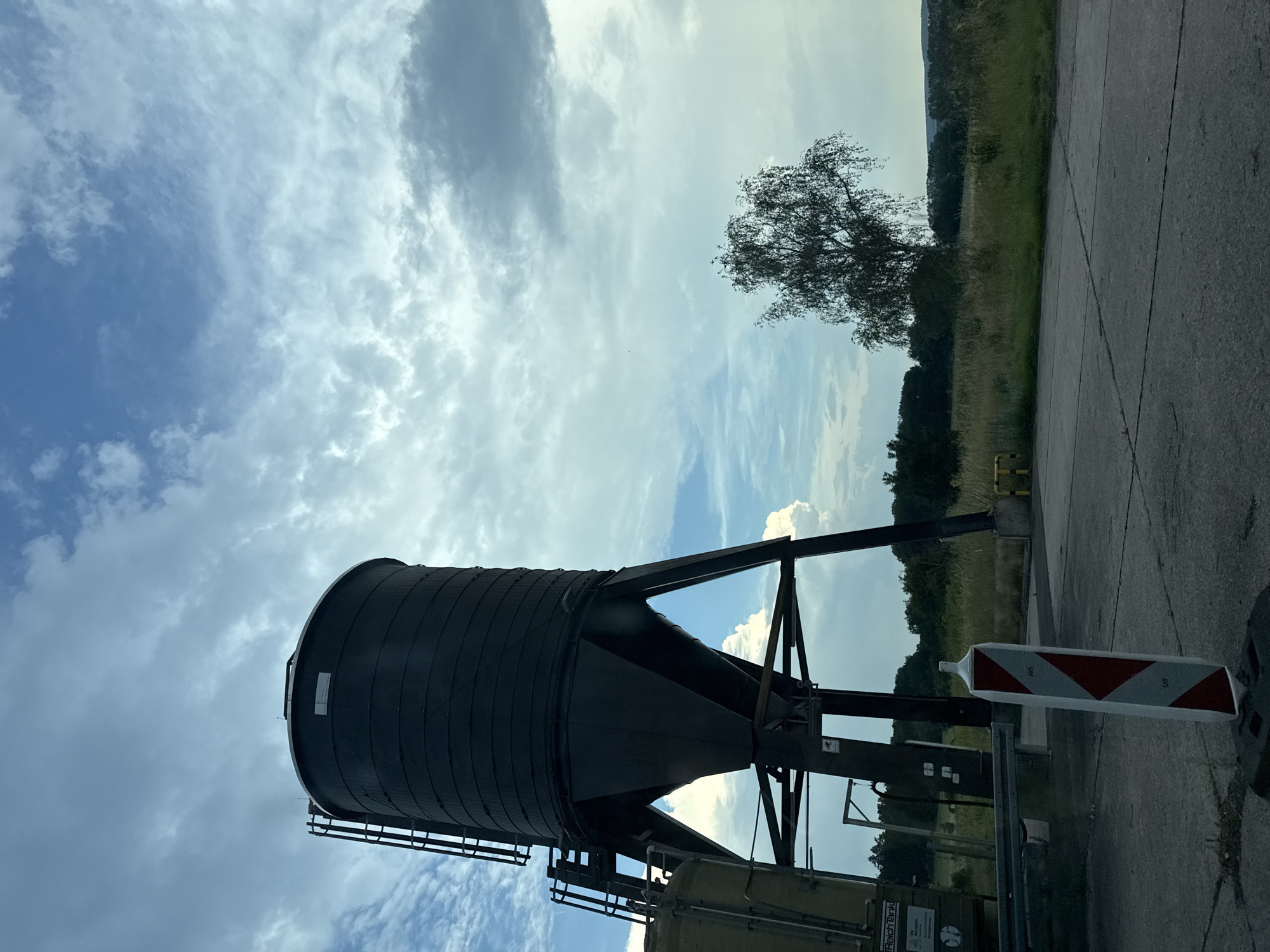 In the city of Linden, Germany, a large, dark silo stands on metal legs against a cloudy sky. To the left of the silo, a portion of another, lighter-colored cylindrical tank is visible. A ladder structure is attached to the side of the dark silo. In the foreground, a striped marker post with red and white sections and the inscription "AVS" stands on a concrete surface. Beyond the silo, a field of tall grass leads to a line of trees, with a single, slender tree with sparse foliage standing out in the middle distance. The sky is filled with a mix of light and dark clouds.