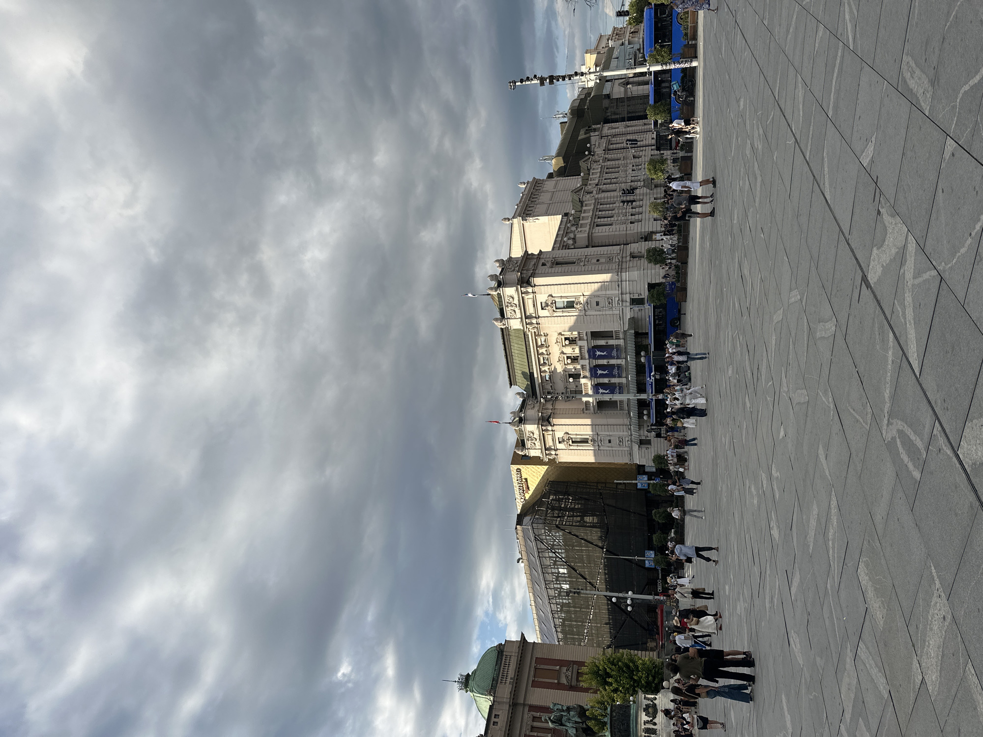 The image depicts a wide, open plaza paved with large, rectangular stone tiles, some of which have a patterned inlay. In the background, a grand, pale-colored building with classical architectural features, including columns and ornate carvings, dominates the scene. To the left of this building, another structure with a green domed roof is visible, adorned with a spire. Adjacent to the classical building, a modern, glass-encased structure stands out, its dark facade contrasting with the older architecture. Several blue buses are parked along the edge of the plaza, with people milling about. A crowd of individuals are scattered across the plaza, some walking, some standing in groups, engaged in conversations. The sky above is filled with dramatic, dark, and light grey clouds, suggesting an overcast or impending weather change. The overall impression is a busy public space in an urban environment. This is Nikola Tesla Square in Belgrade, Serbia.