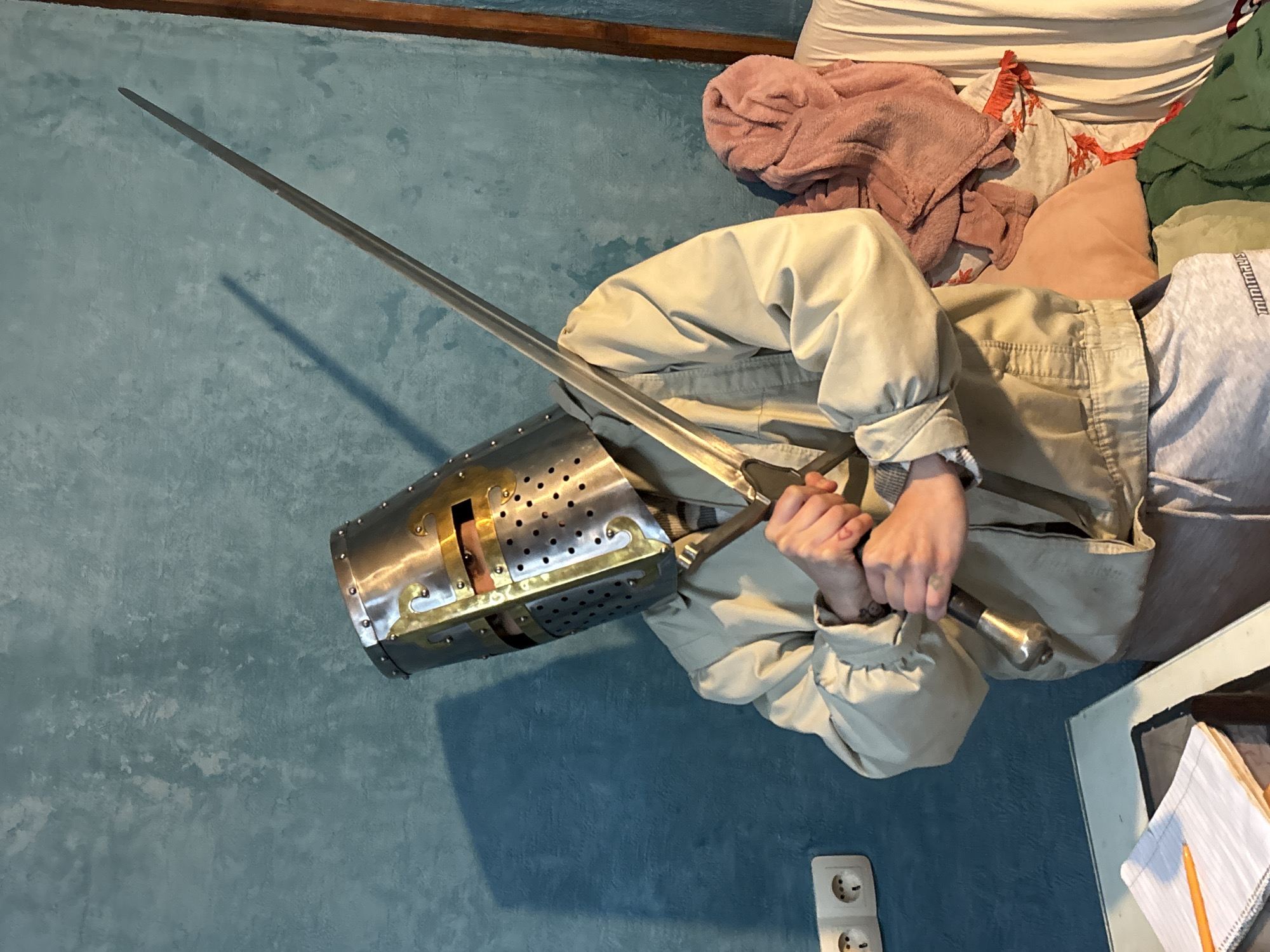 A person wearing a Crusader helmet and a tan jacket holds a sword. They are on a bed with pillows and blankets, with a notebook visible on a table nearby. The background is a blue wall with a European-style electrical outlet. The location is unknown; the suggestion of Verinsko, Bulgaria is not confirmed by the image.
