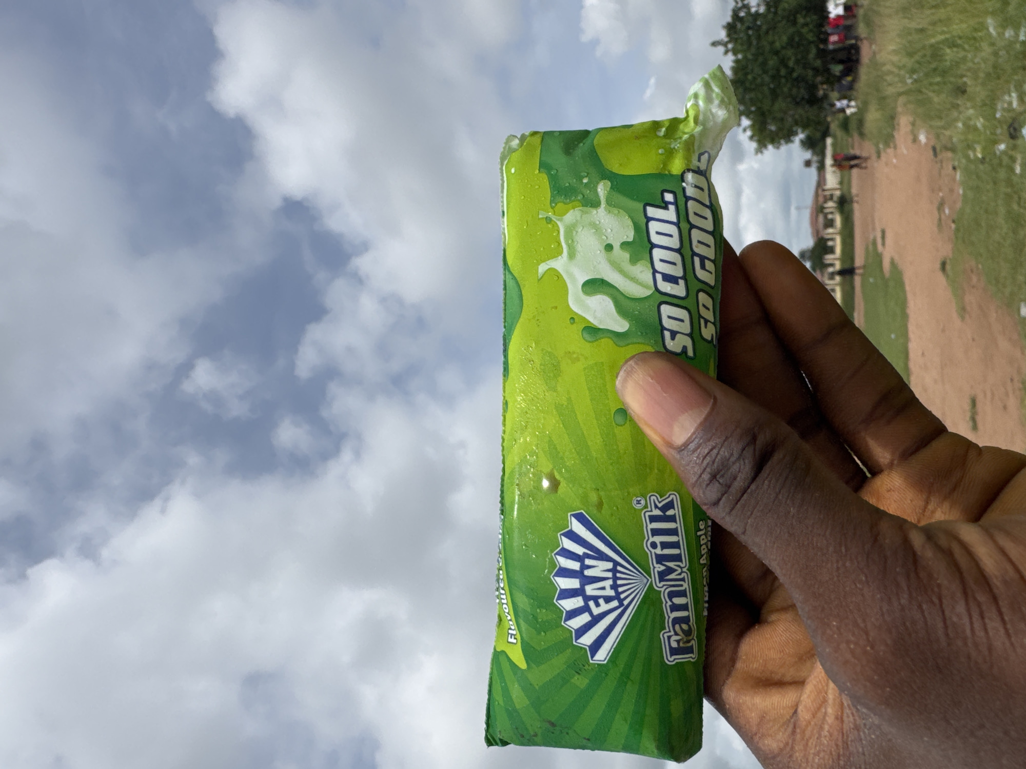A person's dark-skinned hand holds a green and white "Fan Milk So Cool" ice cream package vertically in the foreground. The package is partially unwrapped at the top. In the background, a cloudy sky fills the left side, while a grassy field with a dirt path and scattered trees occupies the right side. The scene appears to be outdoors. The location is Ihiagwa, Nigeria. A few indistinct figures are visible in the background, possibly walking on the path.