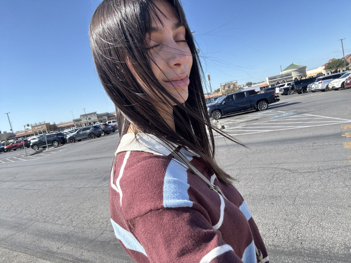 A woman with long, dark hair stands in a parking lot with her eyes closed, seemingly enjoying the sun. She wears a patterned burgundy and light blue sweater with a light-colored collar. The parking lot is filled with cars of various makes and models. A shopping center is visible in the background under a clear, blue sky.