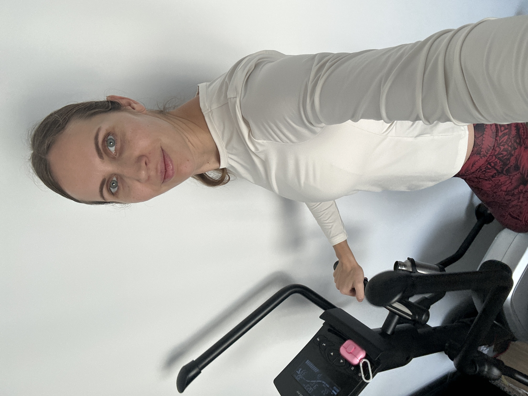 A woman is standing next to an elliptical machine. She has light brown hair pulled back and is looking at the camera. She is wearing a long-sleeved white shirt and red leggings. Her right hand is on one of the elliptical handles. The elliptical machine has a digital display. The background is a plain white wall.