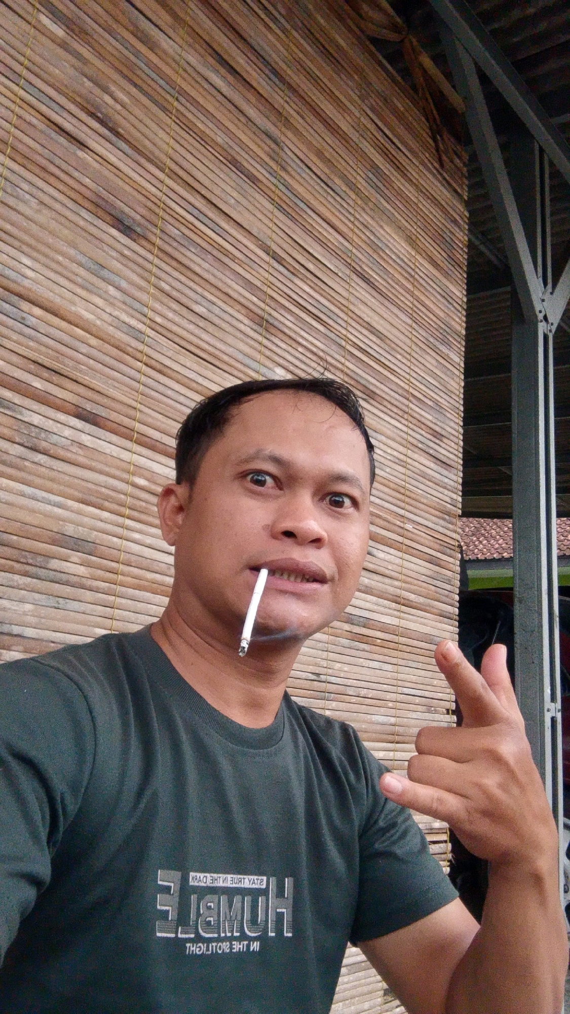 A medium shot captures a man in his late 30s, of Asian descent, holding a cigarette in his mouth and making a rock-on hand gesture. He is wearing a dark green t-shirt with a reflective white design that reads "HUMBLE" in large block letters and "STAY IN THE TRUE DARK" and "IN THE SPOTLIGHT" in smaller, reversed text above and below. The man has short, dark hair and is looking directly at the camera with a playful expression.
The background consists of a woven bamboo wall, likely a blind or screen, and a portion of a metal roof structure with a dark gray beam. The lighting suggests it is daytime, possibly outdoors or in a covered patio area. The scene appears to be in Cintaratu, Indonesia, a coastal village in the Tasikmalaya Regency, West Java.