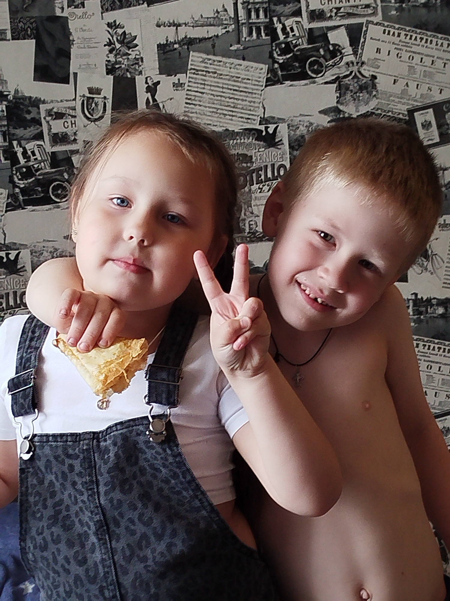 Two young children, a girl and a boy, are posing for a photo in front of a wall decorated with a collage of vintage advertisements and images. The girl, on the left, has blonde hair and blue eyes, and she is wearing a white top with dark denim overalls. Her arm is around the boy's neck, and she is holding a piece of food, possibly a pastry or pancake, in her hand. The boy, on the right, has light brown hair and is shirtless, wearing a dark necklace with a cross pendant. He is smiling broadly and holding up two fingers in a peace sign. Both children appear to be happy and playful. The background wallpaper features various vintage graphics, including images of old cars, theatrical posters, and travel-related designs. There is no clear indication of the time of day or weather, but the indoor setting suggests it's a casual moment at home. Visible text on the wallpaper includes "RIGOLETTO," "FAUST," "CHIANTI," and "VENICE." The location context provided is Chaikovsky, Russia.