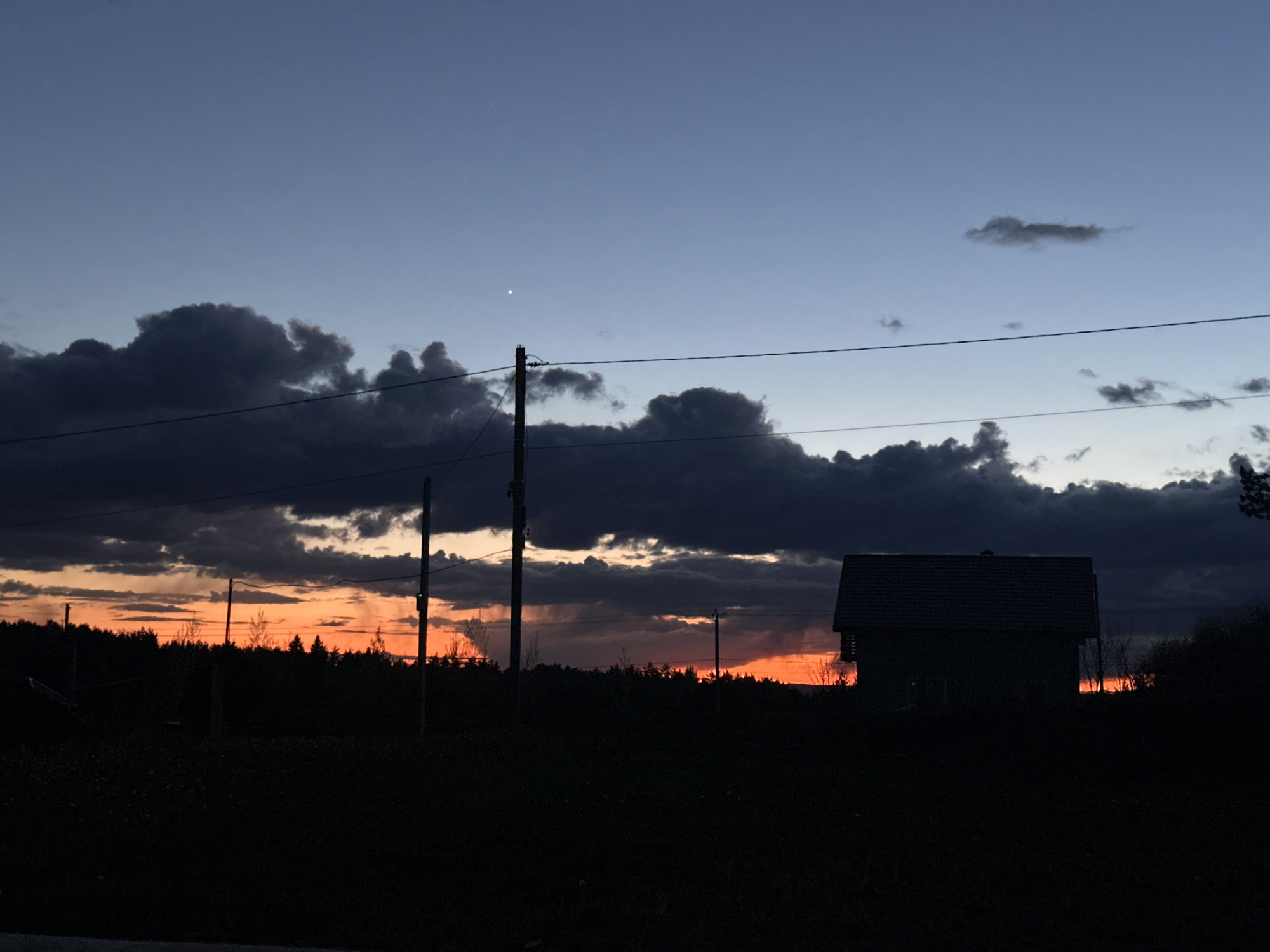Captured at dusk or dawn in the city of Прикамский, Russia, the image showcases a silhouetted landscape dominated by the silhouette of a house on the right. The sky, a blend of blue and orange hues, is partially obscured by dark, ominous clouds. Power lines stretch across the upper half of the frame, supported by utility poles. The foreground is entirely black, obscuring any details of the terrain. Trees are visible in the distance, their forms vague and indistinguishable against the skyline. A single bright star or planet is visible in the sky.