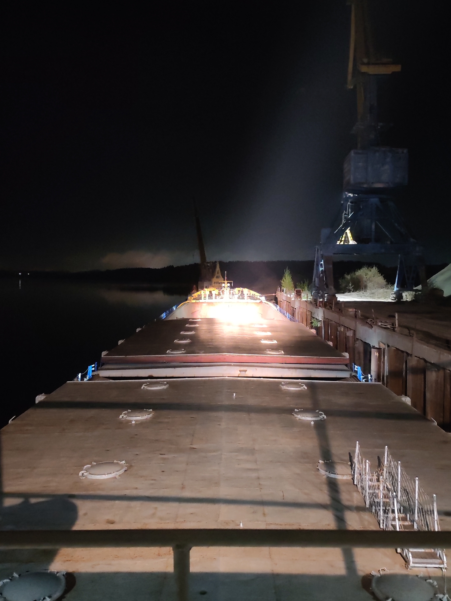 At night, a large barge is docked at the Port of Tver on the Volga River in Tver, Russia. The barge’s deck is visible, featuring several circular hatches and a ladder. In the background, a large industrial crane is positioned on the dock. The river extends into the distance, and the opposite bank is lined with trees. The sky is dark, illuminated by the lights on the barge and dock.