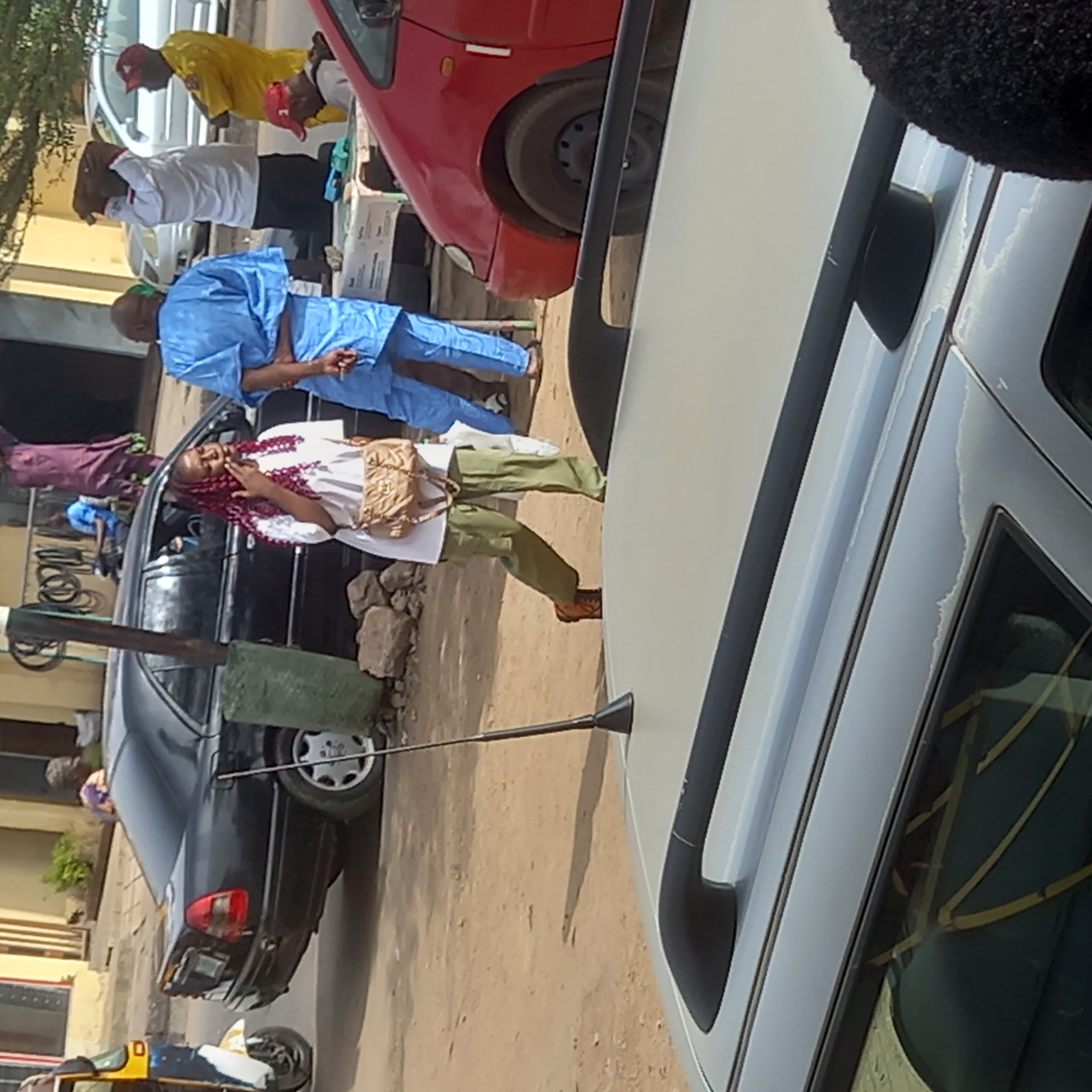 The image, taken in the city of Sardauna, Nigeria, depicts a street scene with several people and vehicles. A woman wearing a white top, green pants, and a tan-colored purse walks on a dirt road. Beside her is a man dressed in a light blue traditional outfit. In the background, three men are standing near a red car, all bowing forward. One man wears yellow, another wears white, and the third is wearing black. A black car is parked beside them. A white car is in the foreground. A three-wheeled vehicle is visible to the bottom left. The environment appears to be a dusty street with buildings visible in the background.