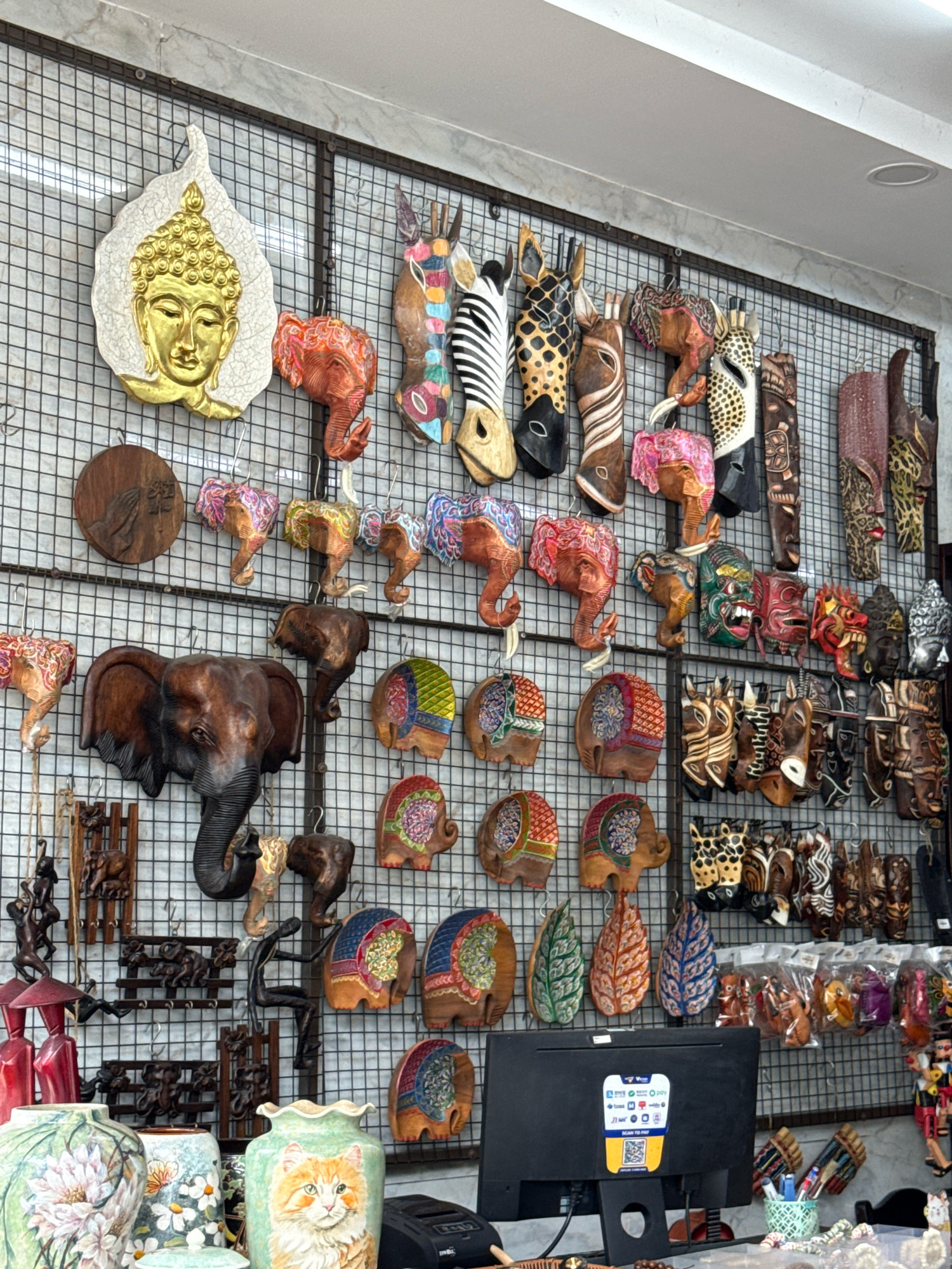 The image depicts a vibrant display of handcrafted souvenirs and decorations, likely within a shop or market stall in Nha Trang, Vietnam. The wall is covered with a metal grid onto which various items are hung and displayed. Dominating the upper left is a large, white, leaf-shaped plaque featuring a golden, relief-carved depiction of Buddha's face.
To the right of the Buddha plaque, a collection of carved wooden animal heads, including elephants and zebras with intricate patterns and bright colors, are mounted. Further to the right, a series of stylized wooden masks, some with fierce expressions and others more abstract, are displayed. These masks exhibit a range of colors and intricate carving details.
Below these, the wall is further adorned with smaller wooden carvings, including more elephant heads, some painted with colorful patterns, and what appear to be carved wooden scenes or figures. Several colorful, leaf-shaped wooden pieces are also visible.
In the lower section of the image, a large, dark wooden carving of an elephant's head with prominent tusks is prominently displayed. To the right of this, a collection of smaller wooden animal carvings and masks are arranged. Rows of small, bagged items, possibly trinkets or further souvenirs, are also visible.
At the bottom of the frame, a black computer monitor is positioned on a counter, displaying a QR code for payment. In front of the monitor, a light green vase painted with a cat's face is visible, alongside other vases and decorative items. The overall impression is one of a colorful and eclectic collection of artisanal crafts.