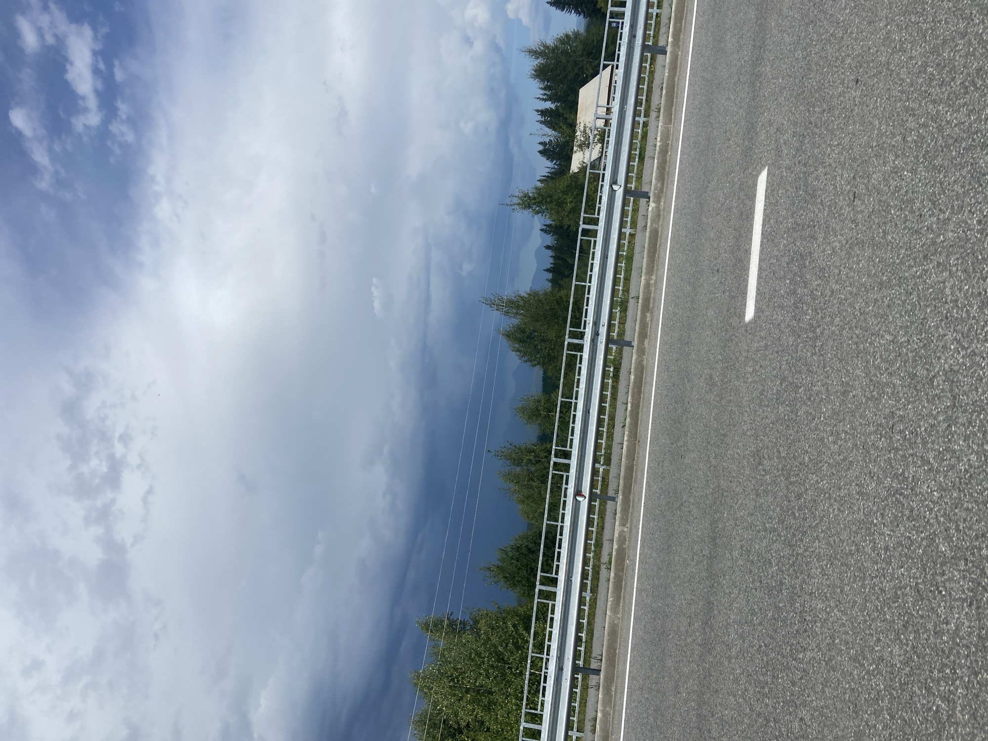 The image displays a highway in Zaktui, Russia, under a cloudy sky. The foreground shows the asphalt of the road with a white line marker. A metal guardrail runs along the side of the road, separating it from a grassy area and a line of trees. In the distance, beyond the trees, a house with a red roof is visible, and a dark, stormy cloud formation looms in the sky. The overall scene suggests a journey or a moment captured during travel.