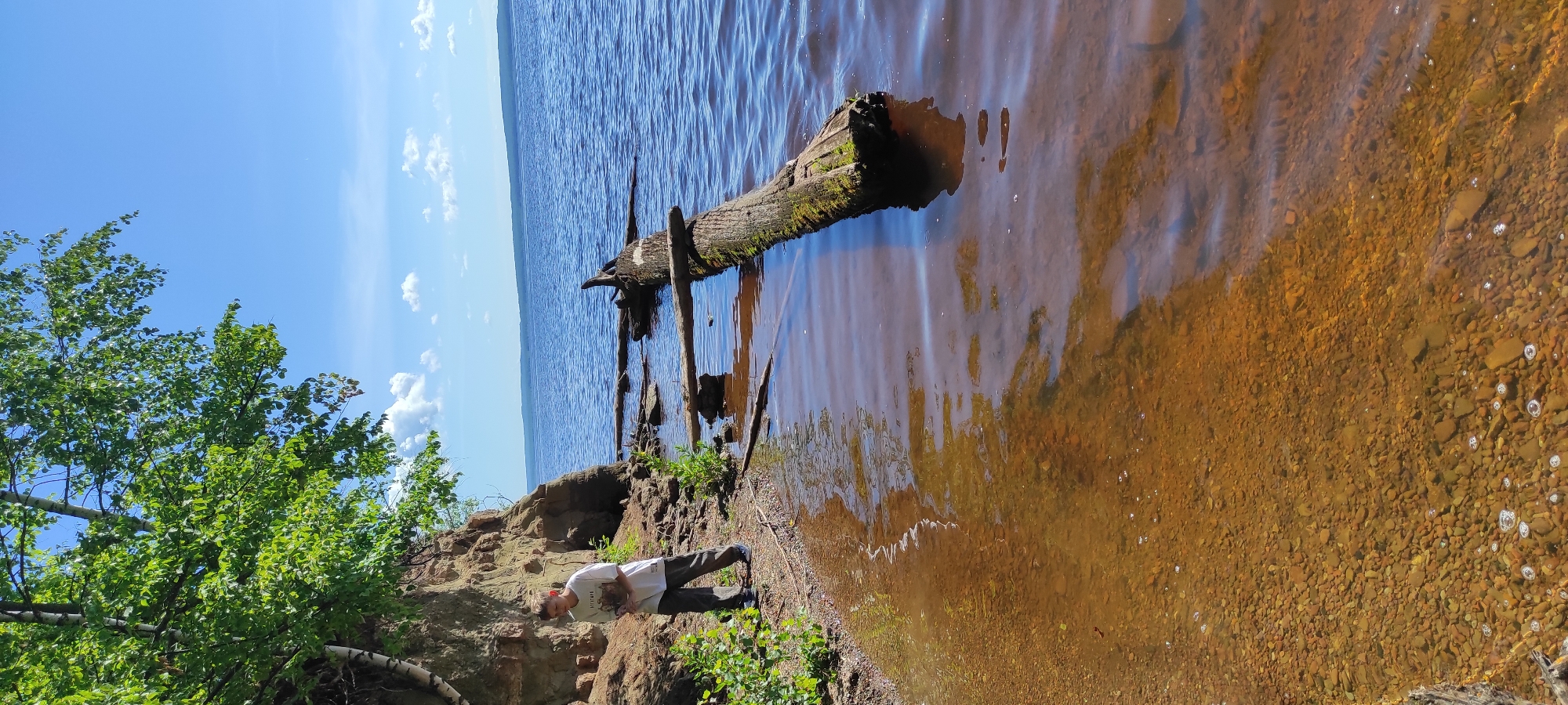 A young boy stands on a pebbly shoreline next to a steep dirt embankment with sparse green foliage. He is wearing a white t-shirt with a graphic and dark pants. The shoreline leads to a body of water, possibly a river or lake, with a submerged, moss-covered log protruding from the water. In the background, a vast blue expanse of water stretches to the horizon, with a distant treeline visible. The sky is a clear blue with a few scattered white clouds. The overall scene depicts a sunny day at a natural waterfront location. The location is likely the Kama River embankment in the city of Chaikovsky, Russia, given the distinctive steep banks and wide river visible in many photographs of the area.