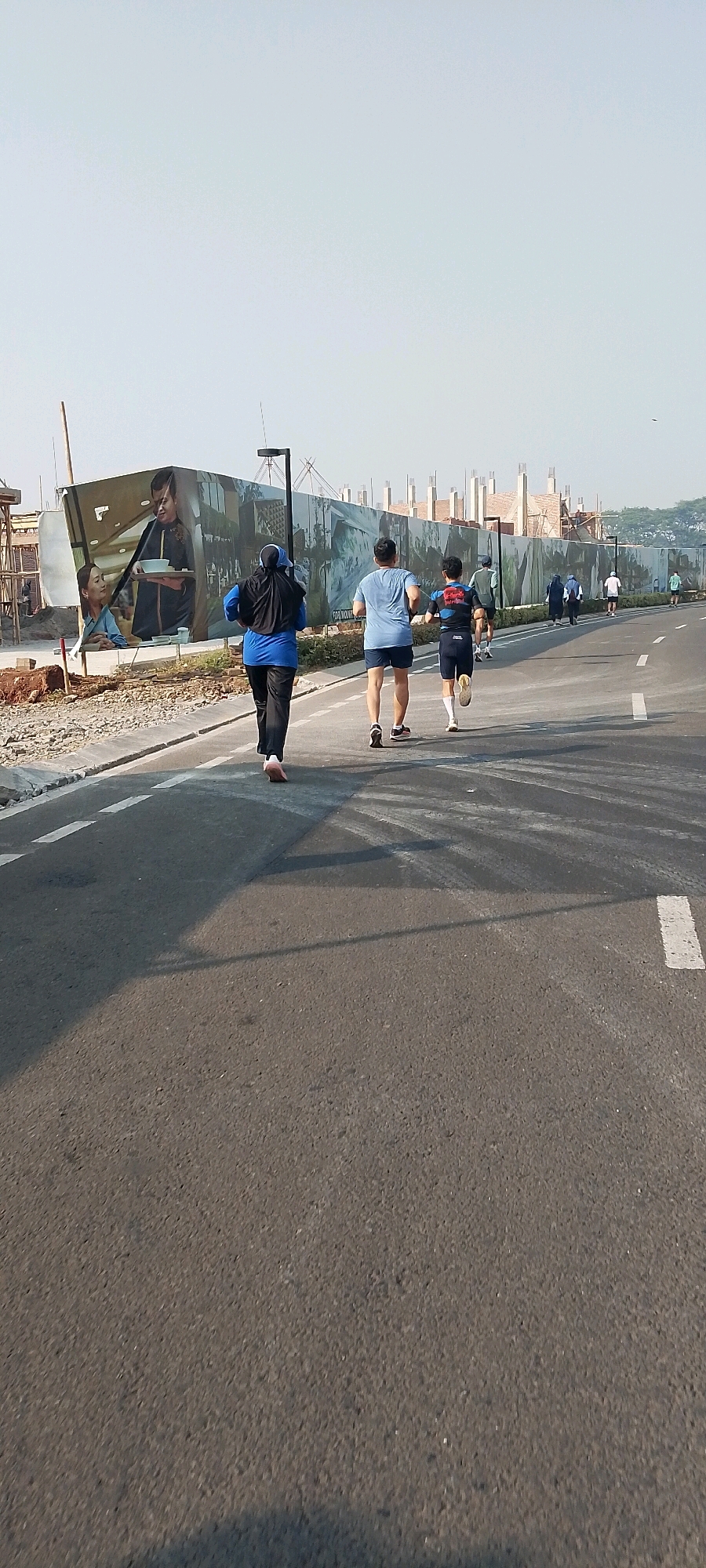 In Gunung Putri, Indonesia, a group of people are jogging along an asphalt road. The image is captured from behind the joggers, showing their backs as they move away from the camera. The road has white dashed lines marking the lanes and a solid white line on the right side. On the left side of the road, a large mural is displayed on a construction hoarding, depicting scenes of people in traditional attire. Behind the hoarding, scaffolding and partially constructed buildings are visible, indicating an ongoing construction site. The sky is clear and bright, suggesting a sunny day. The joggers are dressed in athletic wear, with one person in a blue hijab and blue athletic top and black pants, another in a blue t-shirt and shorts, and a third in a dark t-shirt with colored accents and dark shorts. Several other individuals are further down the road, also jogging or walking.