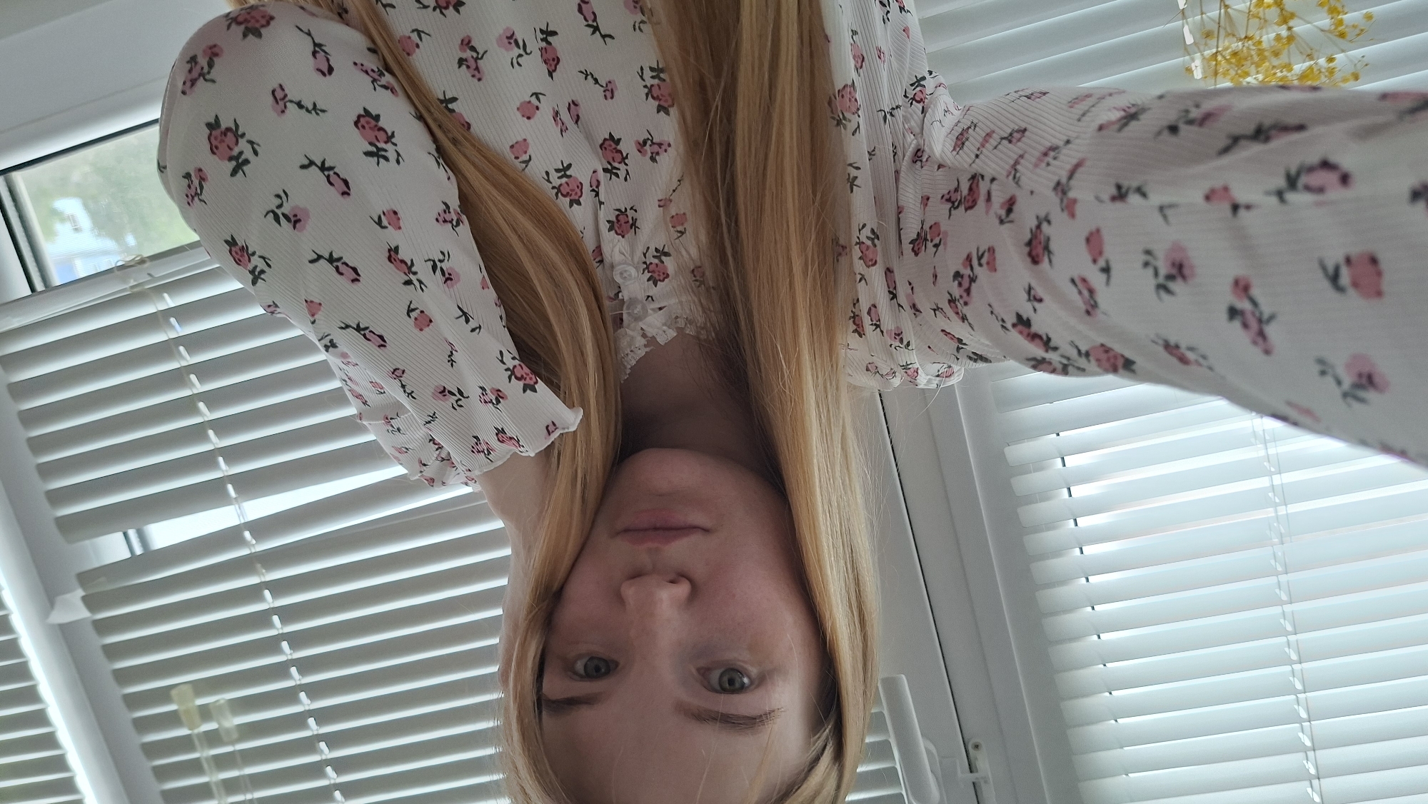 A young woman with long, straight, light blonde hair and a light complexion, exhibiting visible freckles, poses for a selfie indoors. She has green-hazel eyes and a neutral, slightly pouting expression. She is wearing a white long-sleeved blouse patterned with small pink roses and green leaves, featuring a lace-trimmed neckline and small white buttons down the front. Her right hand (viewer's left) extends towards the camera, holding the device, while her left hand (viewer's right) rests against the side of her head. The background consists of a large white window fitted with horizontal white venetian blinds that are partially closed. Through the gaps in the blinds, particularly on the right side, a blurred view of green foliage and a light-colored building or structure suggests an outdoor urban or suburban environment during daylight hours. A white window frame and a white handle are visible. In the lower left portion of the image, a small amount of yellow floral arrangement in a white container is visible. There are no discernible visual markers within the image to confirm the specific location as Chaikovsky, Russia.