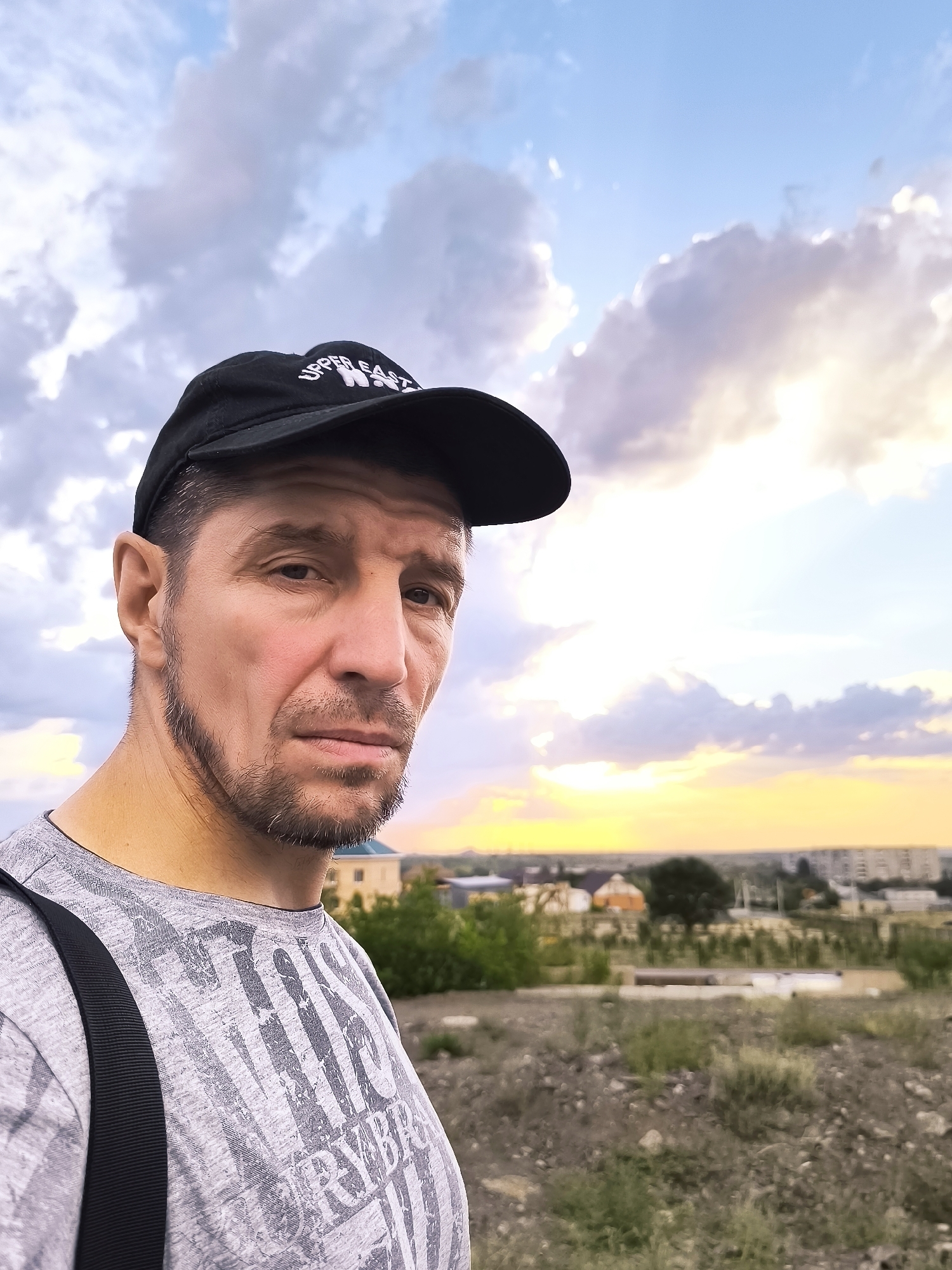 A close-up, selfie-style portrait shows a man wearing a black baseball cap and a grey t-shirt with a printed design. He has short brown hair, a goatee, and is looking directly at the camera with a neutral expression. Behind him, the sky is filled with dramatic clouds tinged with the warm colors of a sunset. In the middle ground, a suburban landscape is visible, with houses, trees, and some construction. The foreground consists of rough terrain with sparse vegetation and gravel. The lighting suggests it is late afternoon or early evening. The scene is set in a semi-arid environment, possibly on the outskirts of a city. There is no indication of the specific location within Qarağandı city administration, Kazakhstan.