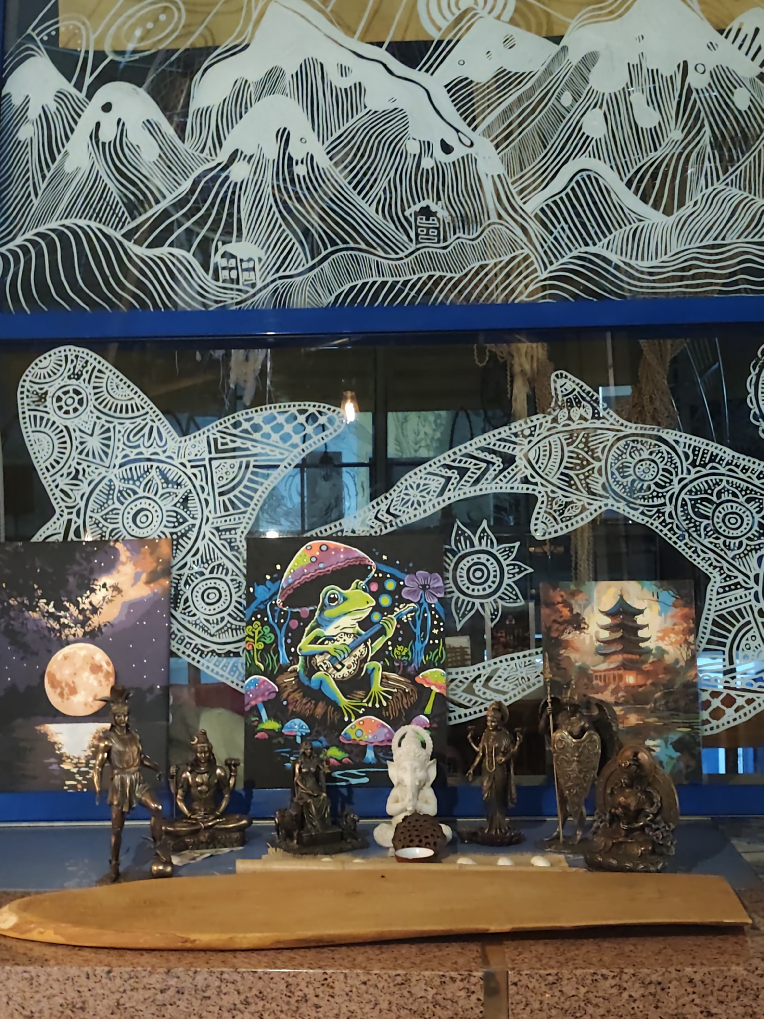 The image depicts an indoor scene, likely a shelf or altar, displaying various artistic and decorative items. Dominating the upper portion of the image is a mural or painting featuring stylized white mountains against a dark background, with delicate, swirling lines and a small structure resembling a house. Below this, a blue-framed window or glass surface is adorned with intricate white line art, forming patterns that suggest celestial bodies or decorative motifs.
In front of the window, several small framed artworks are arranged. On the left, a painting depicts a large, orange full moon rising over a dark landscape with silhouetted trees. To its right, a vibrant, psychedelic-style painting features a green frog sitting under a colorful mushroom, surrounded by other mushrooms and mystical elements. On the far right, another painting shows a pagoda-style building with warm, autumnal colors.
Below these paintings, a collection of small statues is placed. These appear to be figurines of deities or figures from mythology, crafted from dark metal and possibly stone. They are arranged on a blue surface, which could be a shelf or the surface of the display. A prominent wooden object, possibly a platter or a piece of art, lies horizontally in the foreground, obscuring the lower part of the figurines. The background behind the figurines is dark and appears to be a wall or cabinet.
The scene is not identifiable as being located in Sochi, Russia. There are no geographical markers or specific cultural elements that link it to that city.