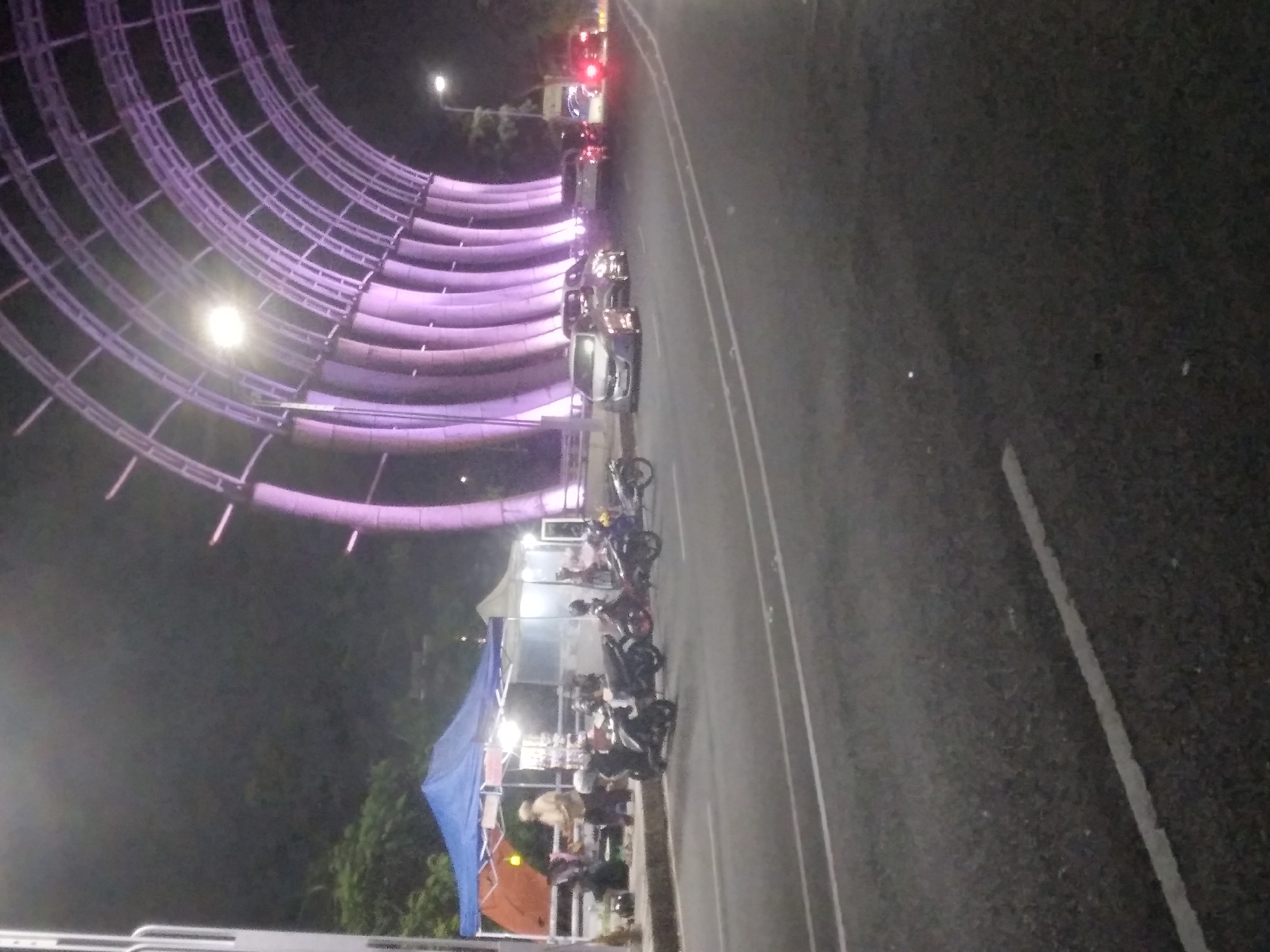 The image depicts a street scene at night in Purwokerto, Indonesia, near the iconic Arc of the City (Gerbang Nam Lim). In the foreground, an asphalt road with white lane markings is visible. To the left, a blue and orange tent is set up, with several people standing and sitting around it, appearing to be vendors or shoppers at a roadside stall. A few motorcycles are parked near the tent.
Dominating the background is the illuminated Arc of the City, a modern archway structure with purple lighting. Several cars are driving on the road, their headlights and taillights visible, indicating nighttime traffic. The overall atmosphere is that of a bustling night market or roadside activity beneath a prominent city landmark.