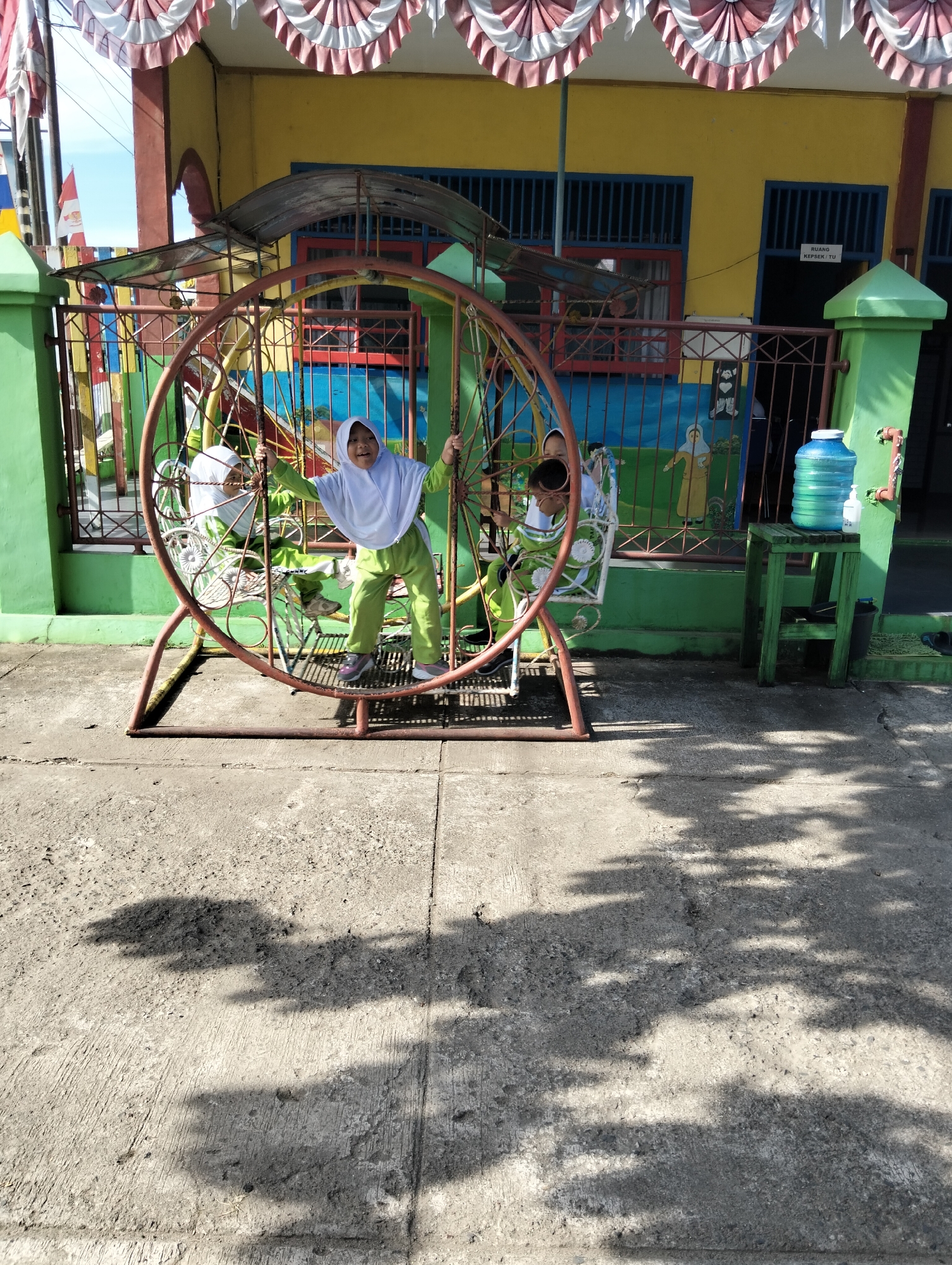 In Janju, Indonesia, at a school, two young girls in hijabs and green uniforms are playing on a large circular swing. The girl in the foreground, on the right side of the swing, is leaning back with her arms extended, smiling and looking at the camera. The other girl, on the left, is also on the swing, partially obscured by the structure. The swing is red and has a circular frame with spokes. The school building in the background is painted yellow and green, with blue-barred windows and a red trim around the doorway. A colorful fence surrounds the play area. To the right of the swing, a green wooden stool holds a large blue water jug and a smaller bottle. The ground is paved with concrete, and shadows of leaves are cast across it, indicating it is a sunny day.