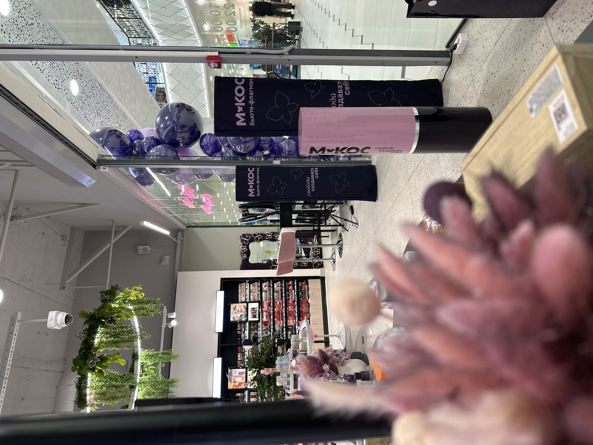 This media file depicts the interior of a modern retail space, likely a beauty or cosmetics store named "MKOC," situated within a shopping mall in Yekaterinburg, Russia. The image is captured from a slightly elevated perspective, looking into the store.
In the mid-ground, at the store's entrance, a vibrant display features numerous purple and light purple balloons. Flanking these balloons are two tall black promotional banners and a large pink cylindrical stand. The banners clearly display the "MKOC" logo, the Cyrillic text "бьюти-флагман" (beauty flagship), and decorative butterfly outlines. The pink stand also prominently features "MKOC" and the slogan "свобода создавать себя" (freedom to create yourself).
Further inside the store, extensive shelves filled with various beauty products, presumably makeup and skincare, are visible. Two light pink bar stools and a mirror with a butterfly-themed frame suggest a customer consultation or testing area. To the left, a circular decorative element adorned with lush green foliage and subtle lighting adds an organic touch to the modern interior.
Beyond the store's glass front, a bright, expansive mall corridor is visible, with two individuals walking in the distance. A sign in the background shows "10:00" and "55:00," likely indicating store operating hours. The flooring throughout is a light-colored speckled tile. In the immediate foreground, a blurred arrangement of dried pink and purple feathery flowers partially obstructs the view, indicating the vantage point is from a display or counter. The scene conveys an active, contemporary retail environment focused on beauty products.