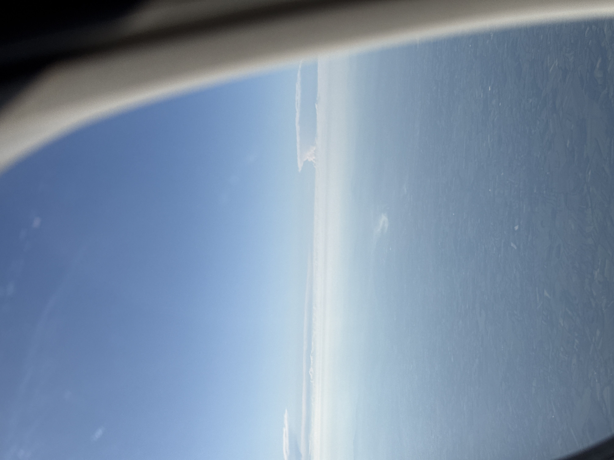 The image appears to be taken from the window of an aircraft. The landscape below is visible, suggesting it is daytime. The sky is predominantly blue with a layer of clouds or haze visible near the horizon. The surface below seems to be a mix of land and possibly water, with the land showing varied textures that could represent fields or forests. The scene includes part of the aircraft window frame at the top. The location in Fiumicino, Italy is not definitively confirmed from the image alone.