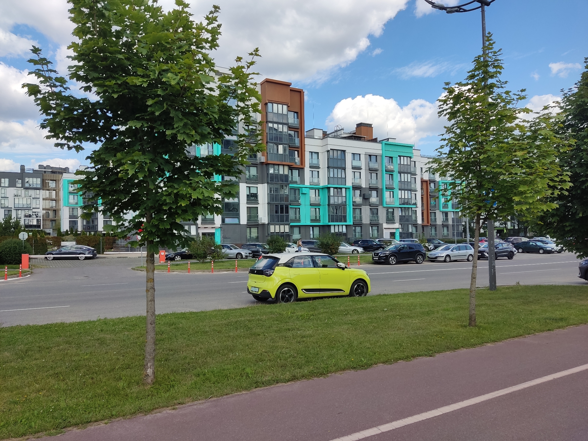 The image captures a bright, daytime scene on a city street, likely in Бараўлянскі сельскі Савет, Belarus. A modern apartment building with a colorful facade, featuring teal, white, grey, and brown sections, dominates the background. Several trees with lush green foliage are positioned in the foreground and midground. A bright yellow compact car is the central focus of the scene, parked on the side of the road. Multiple other vehicles, including sedans and SUVs, are visible on the street and in parking areas. The environment includes a paved road, a grassy median, and a sidewalk or bike path. The sky is blue with scattered white clouds. Orange bollards mark the edge of the road and parking areas.