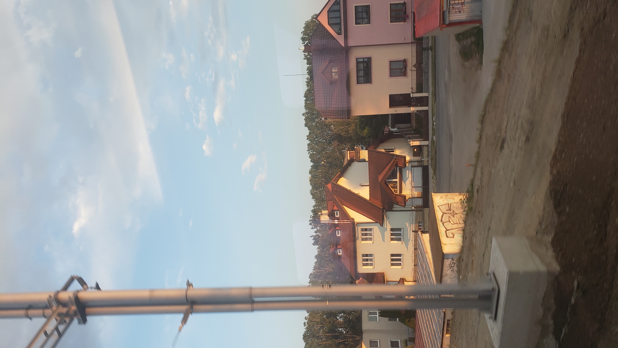 The image shows a street scene in Poprad, Slovakia, with several houses visible. In the foreground, a metal pole with concrete base stands to the left. Beyond the pole, a low concrete structure with graffiti is situated. Further back, a row of houses with red-tiled roofs lines the street. The houses are of varying sizes and architectural styles, with some featuring multiple windows and chimneys. A line of trees forms a backdrop behind the houses. The sky is clear with scattered clouds.